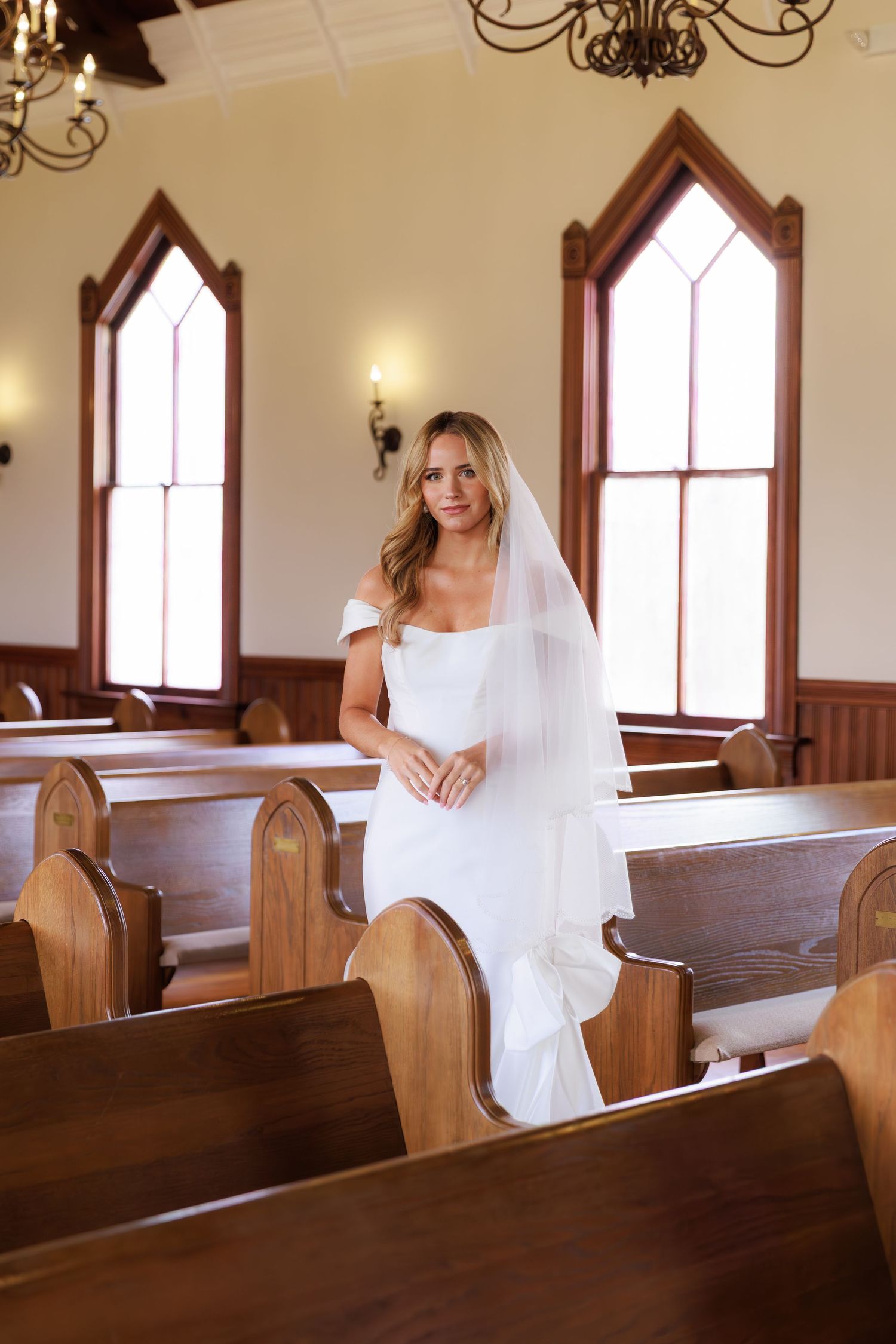 A bride in white poses playfully along church pews in a sunlit chapel with stained glass windows.