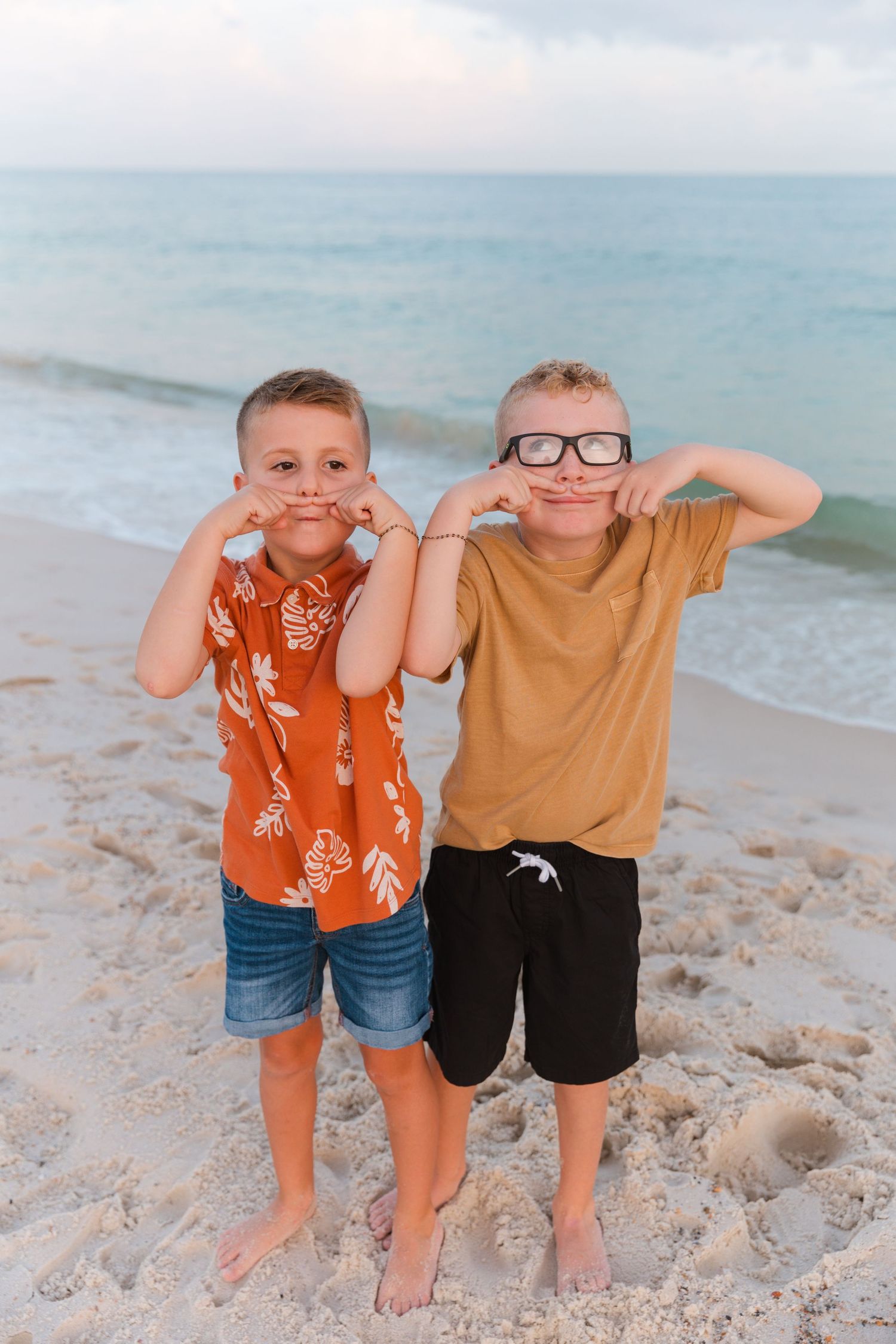 Two children stand together making silly poses on a beach with the ocean behind them.