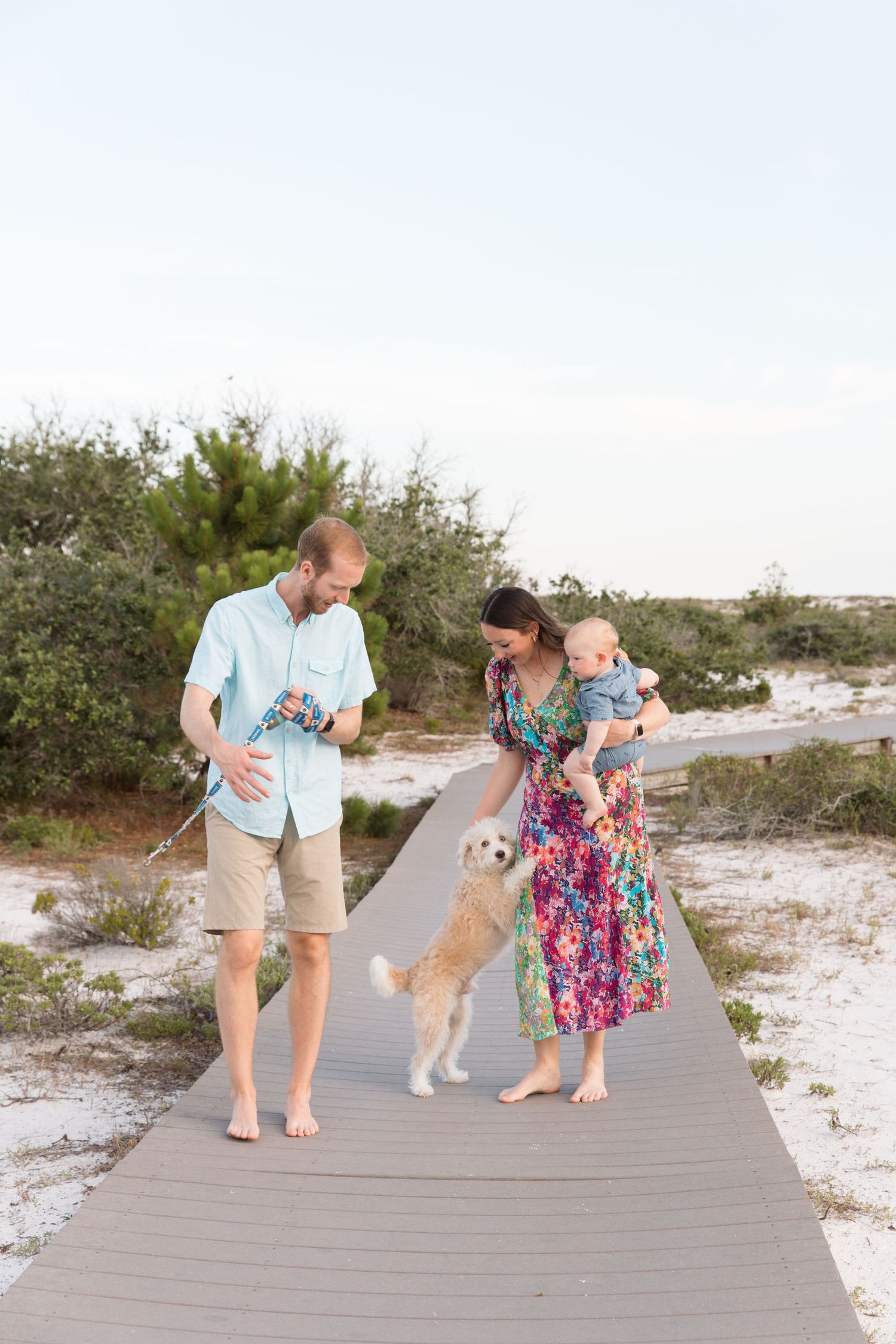 A family walks with their white dog along a wooden boardwalk path near the beach.