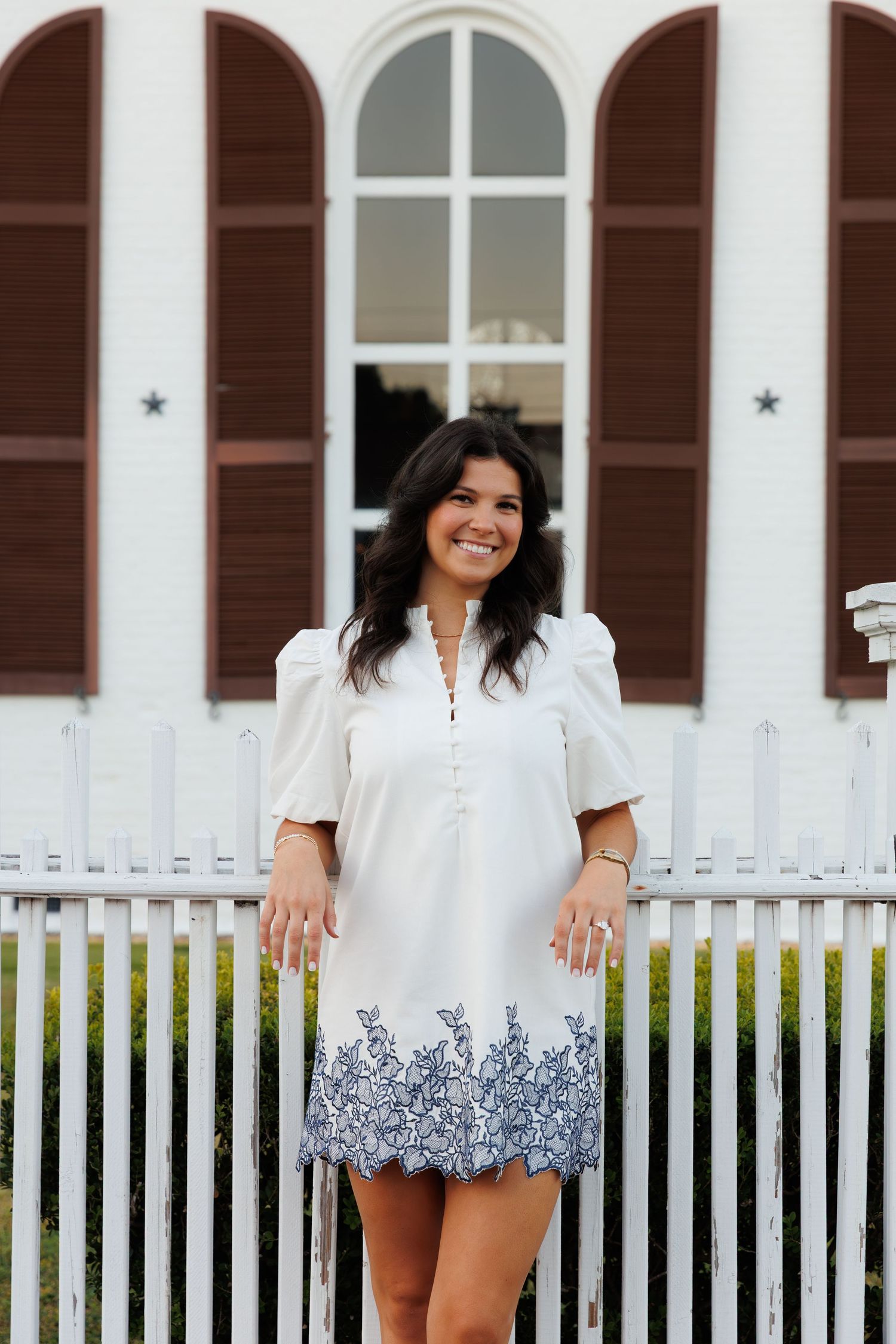 Smiling brunette woman in white embroidered dress poses by white picket fence outside.