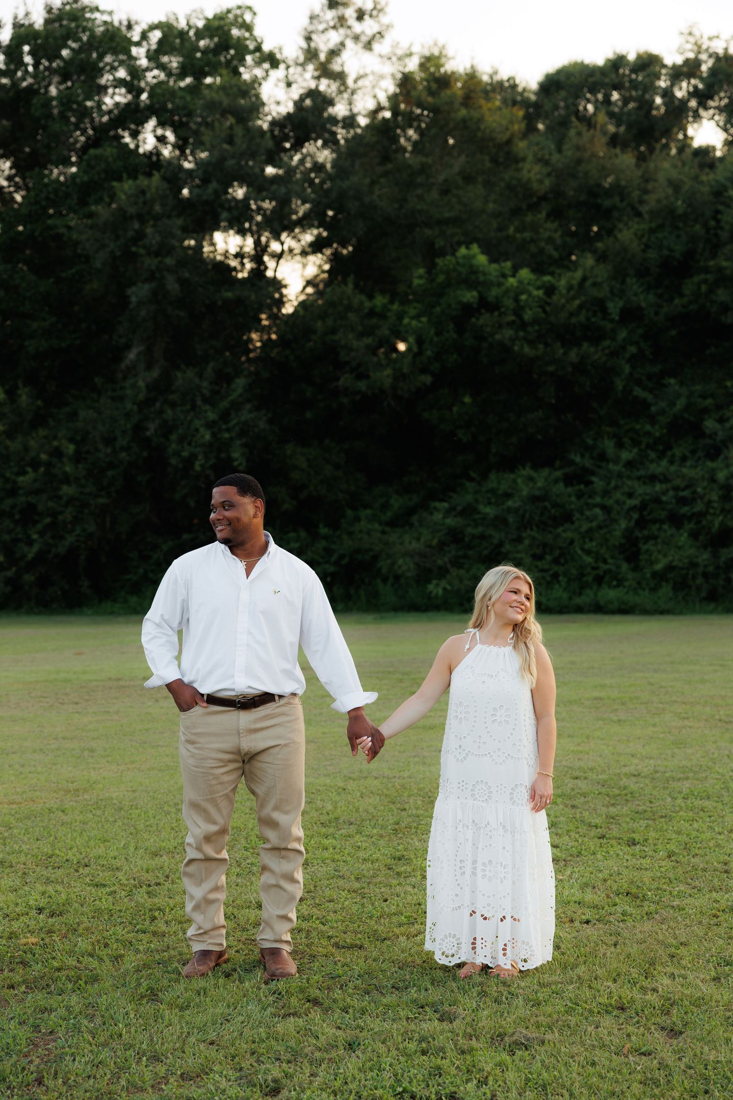 Smiling couple holding hands in a grassy field at sunset, man in white shirt and woman in white dress.