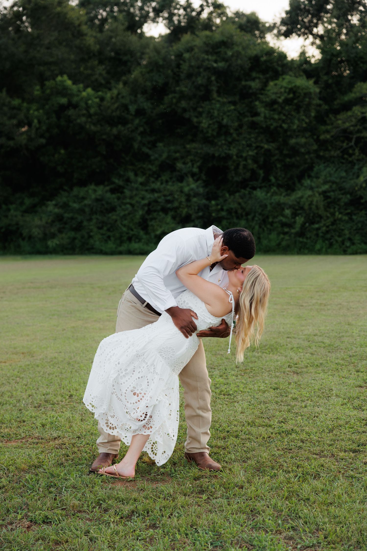 Couple sharing a passionate dip kiss in a lush green field with dense trees in background.