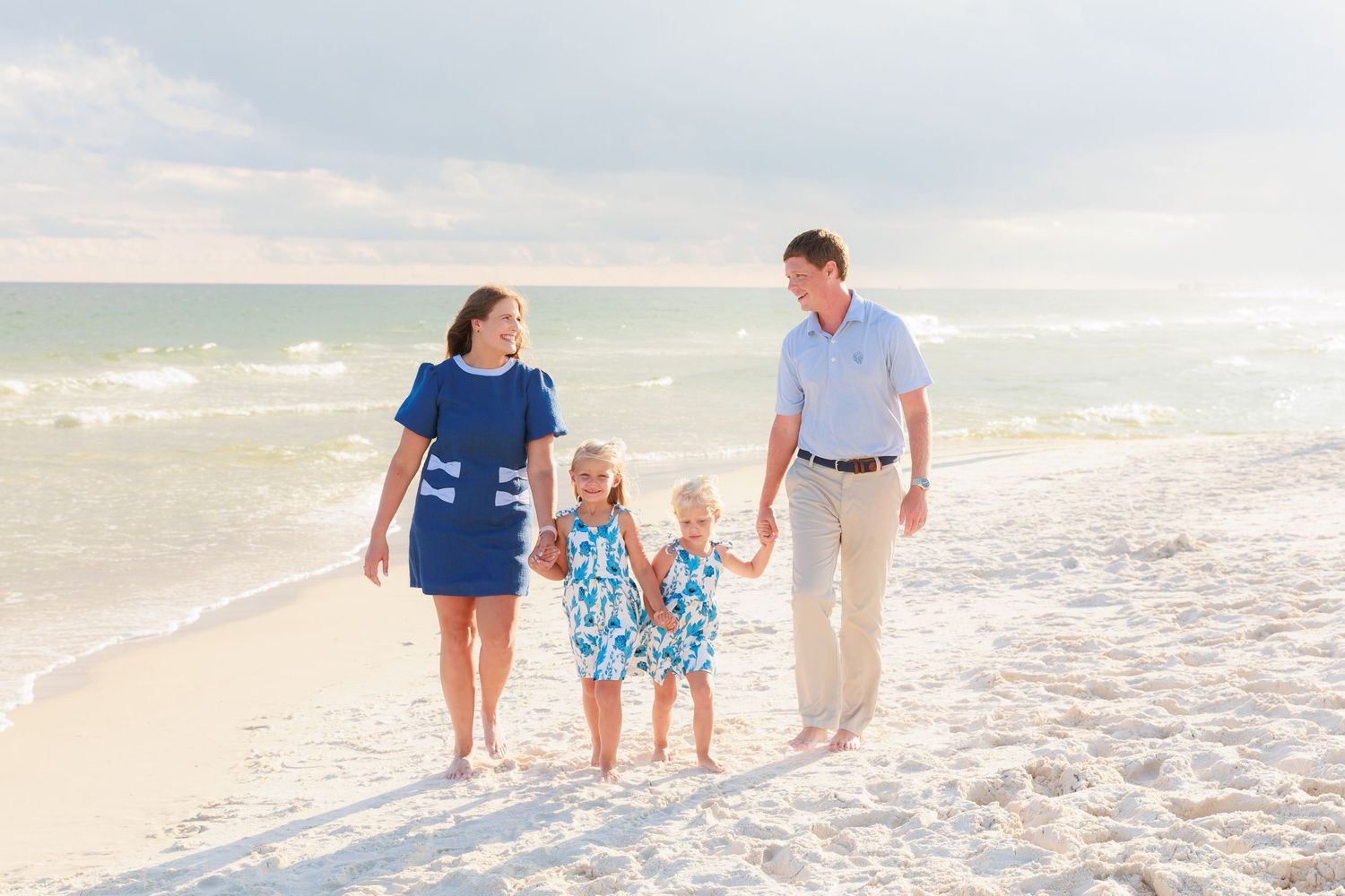 Family of four walking hand-in-hand on a white sandy beach with turquoise ocean waves in the background.