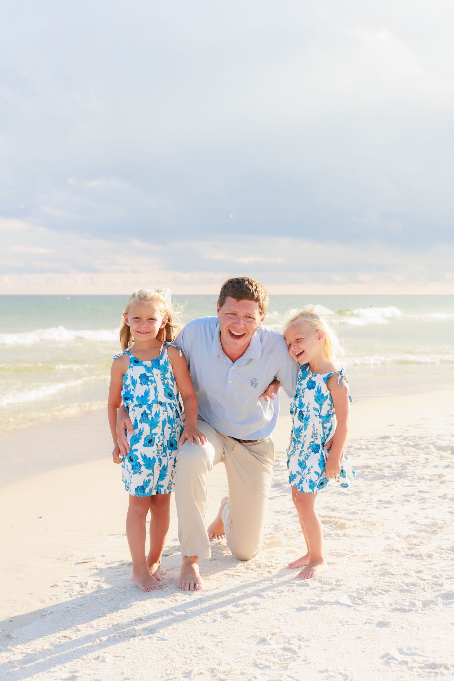 A laughing father kneels on a sandy beach with his two young daughters in matching blue floral dresses near ocean waves.