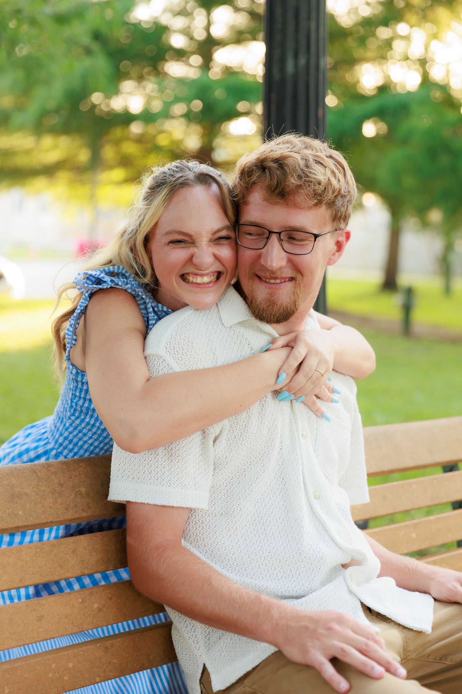 A smiling woman in a blue dress hugs a man with glasses on a park bench outdoors.