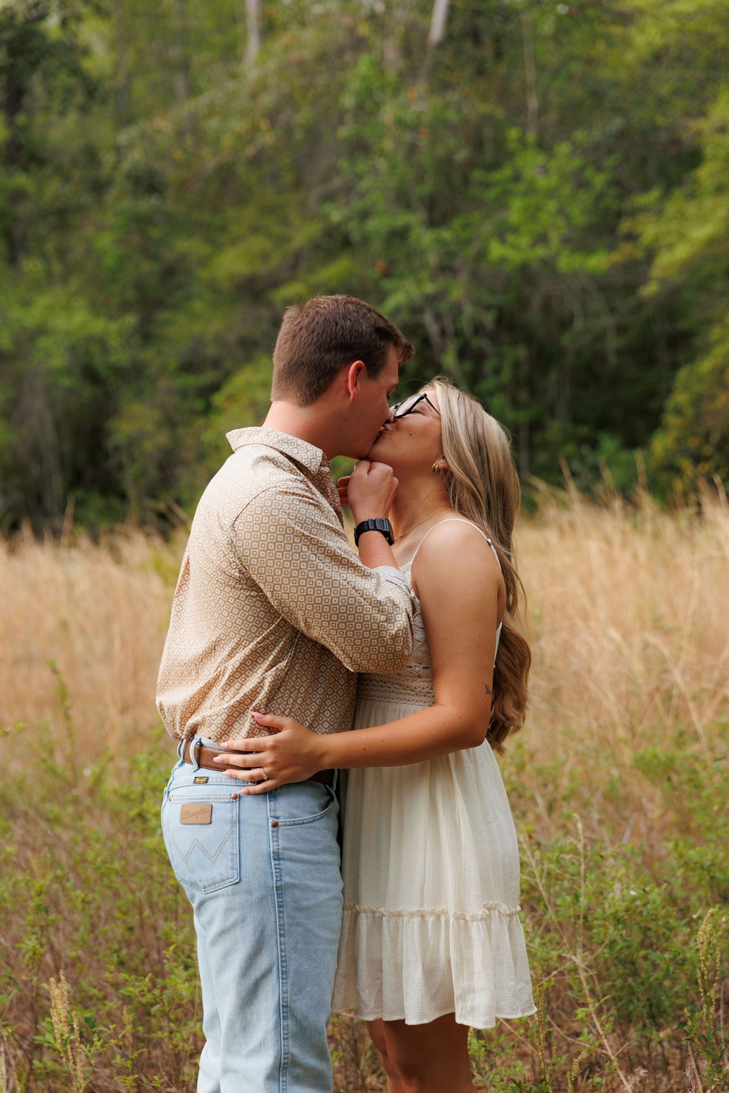 Couple sharing a romantic kiss in a golden field with lush green trees in the background.