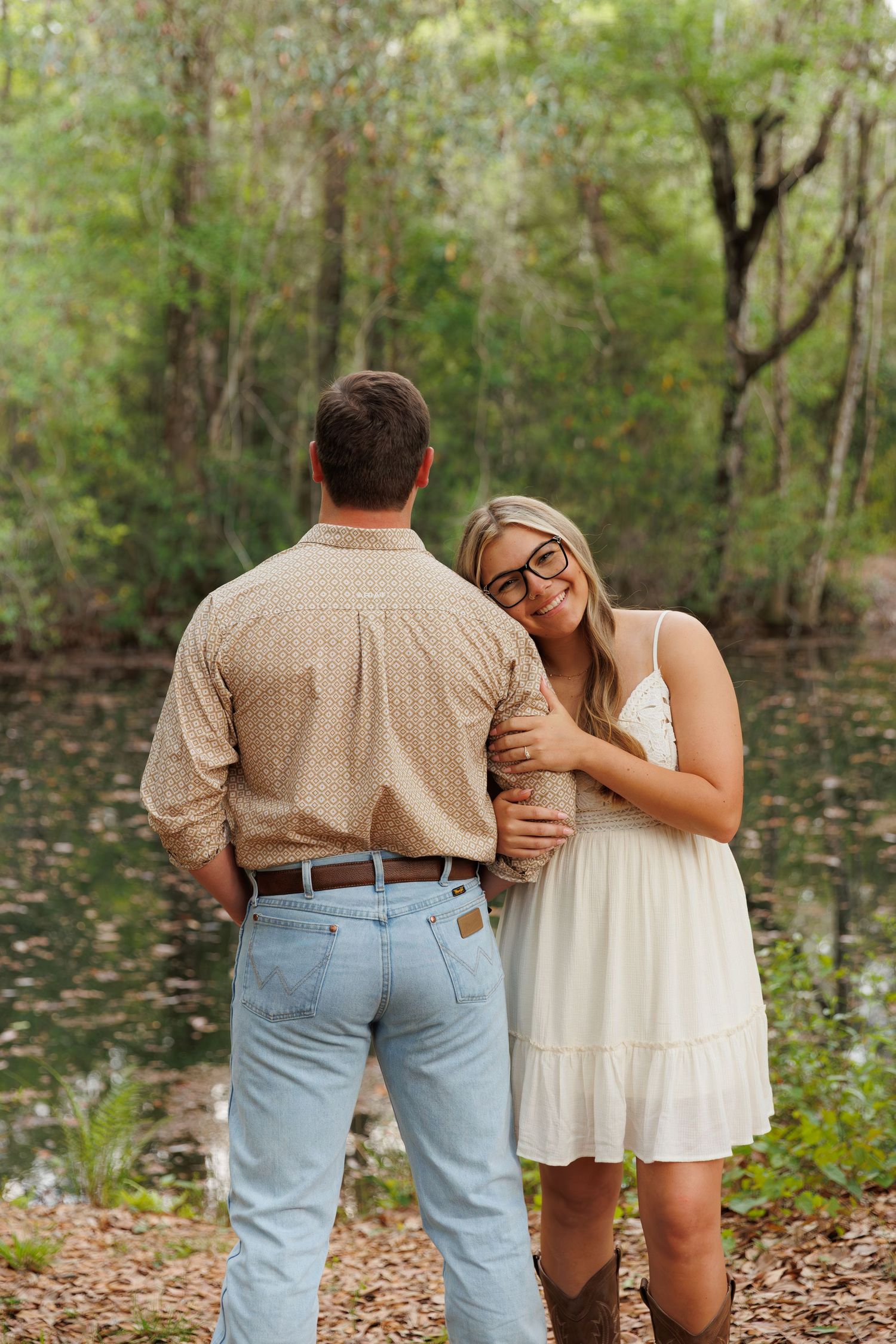 A smiling woman in a white dress and glasses leans on a man in a beige shirt by a forest pond.