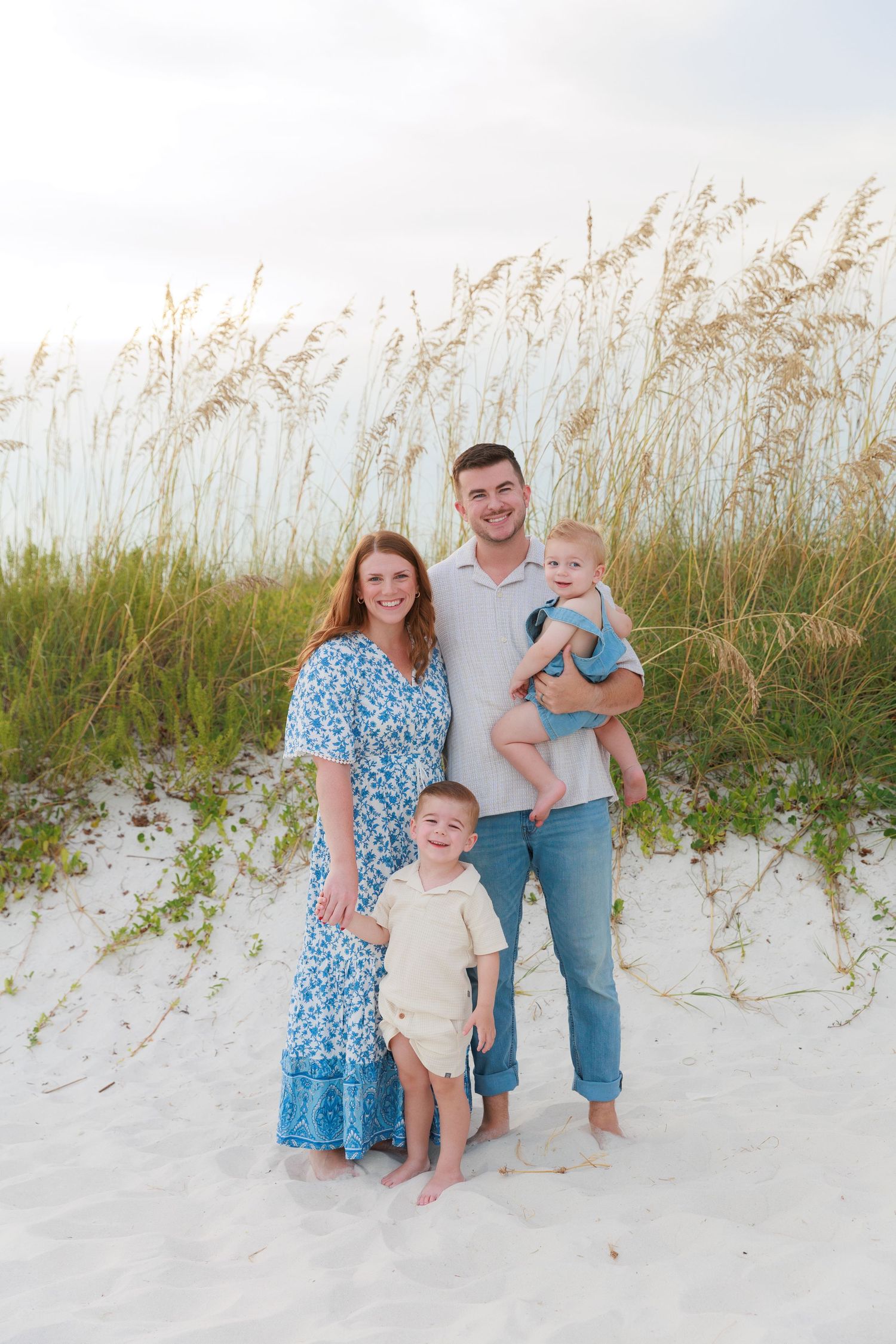 Family of four posing on white sand beach with sea oats, mom in blue floral dress, dad holding baby, toddler standing.