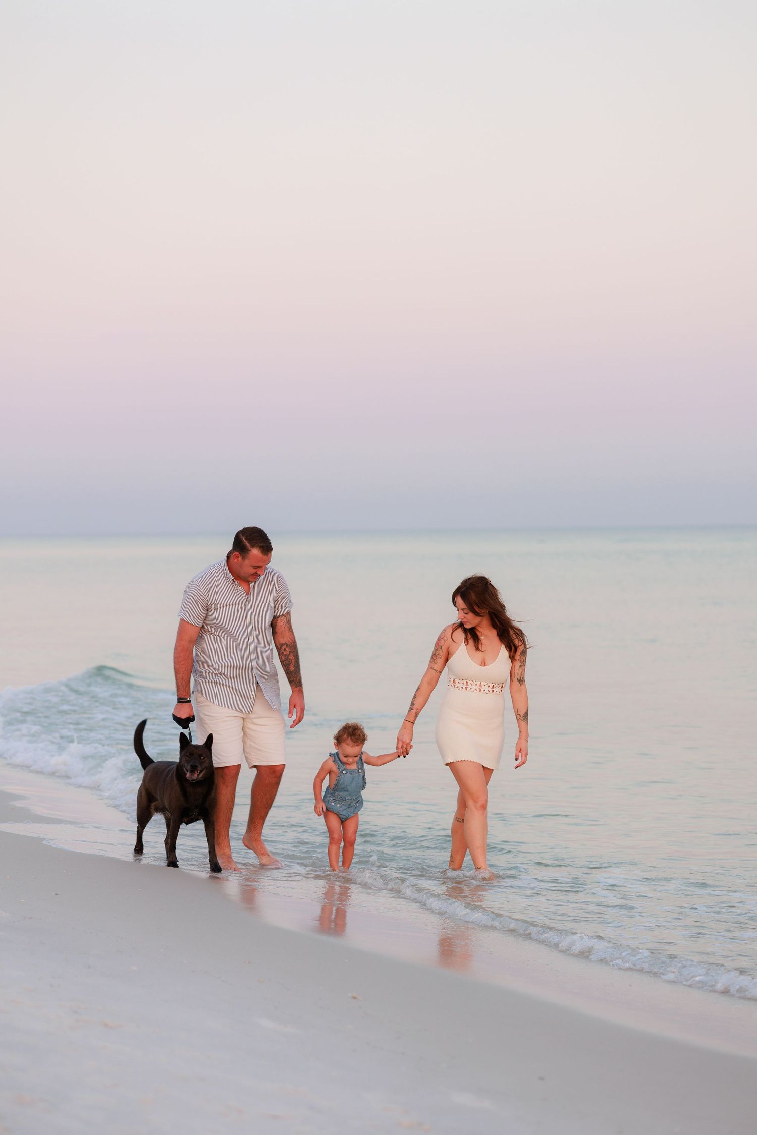 Family with toddler and black dog walking barefoot on a sandy beach at sunset.