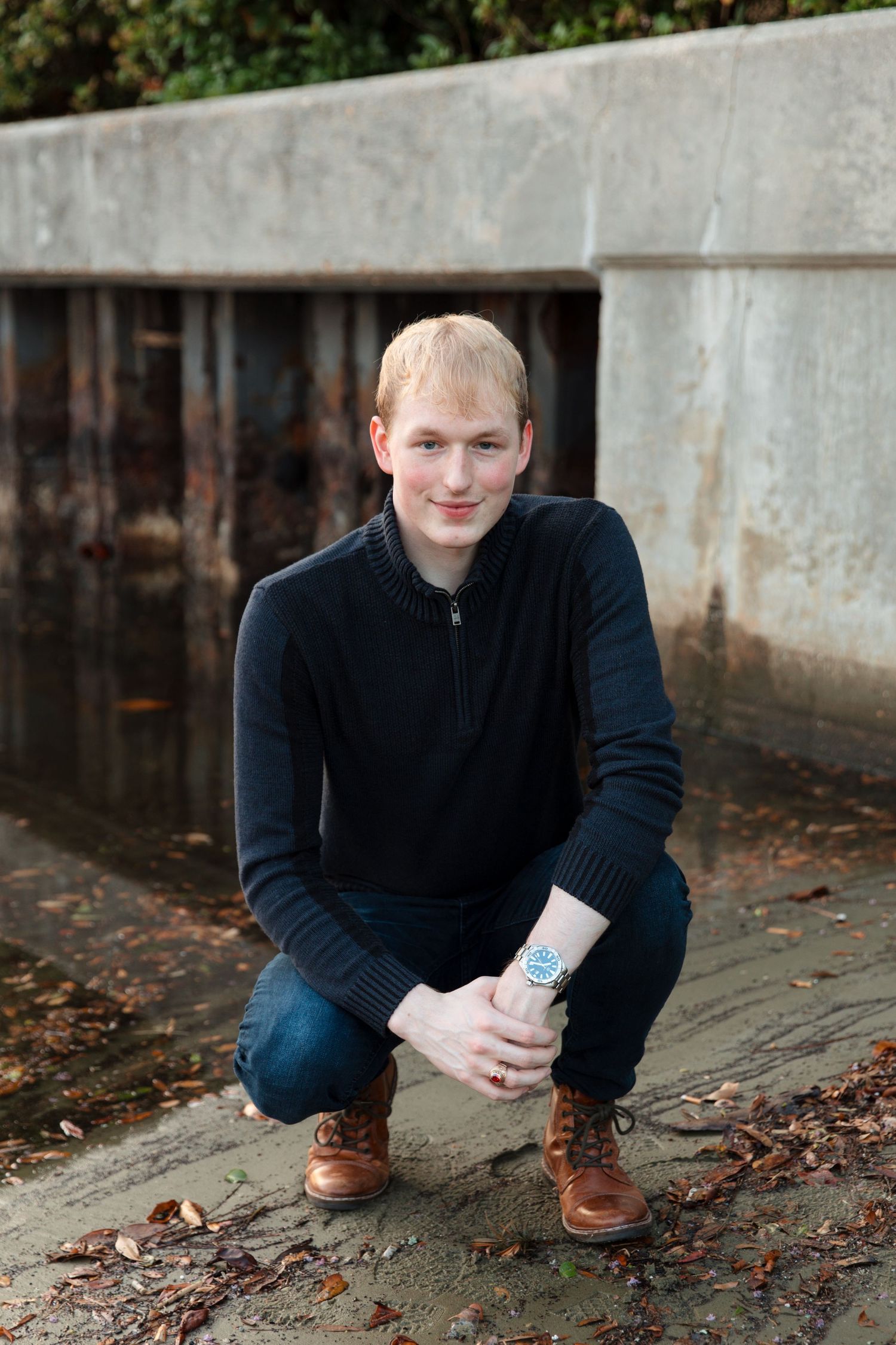 Young blond man crouching in black sweater, jeans, and brown boots outdoors near a concrete wall.