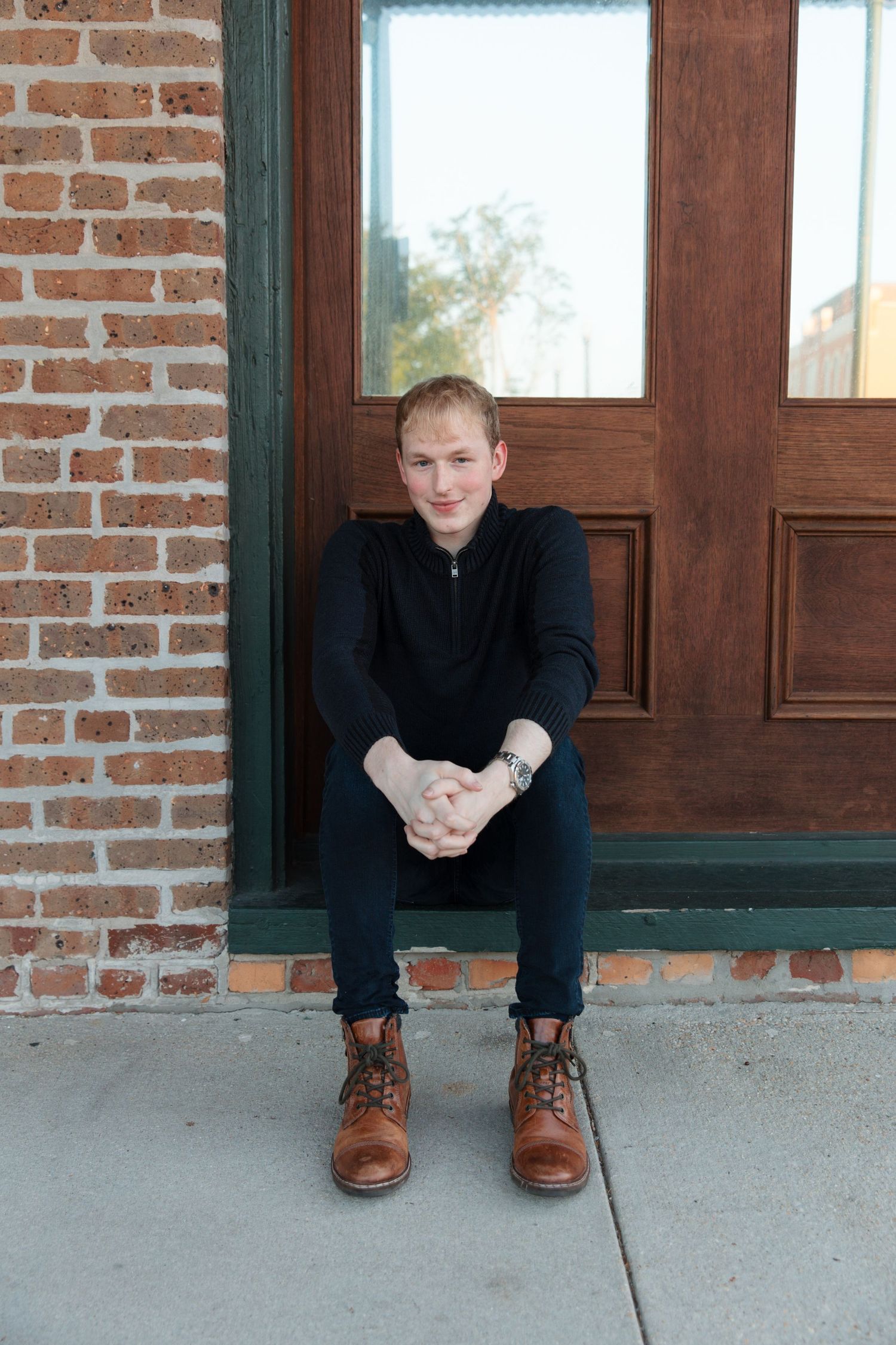 Young man in black outfit and brown boots sitting on steps by a brick wall and wooden door.