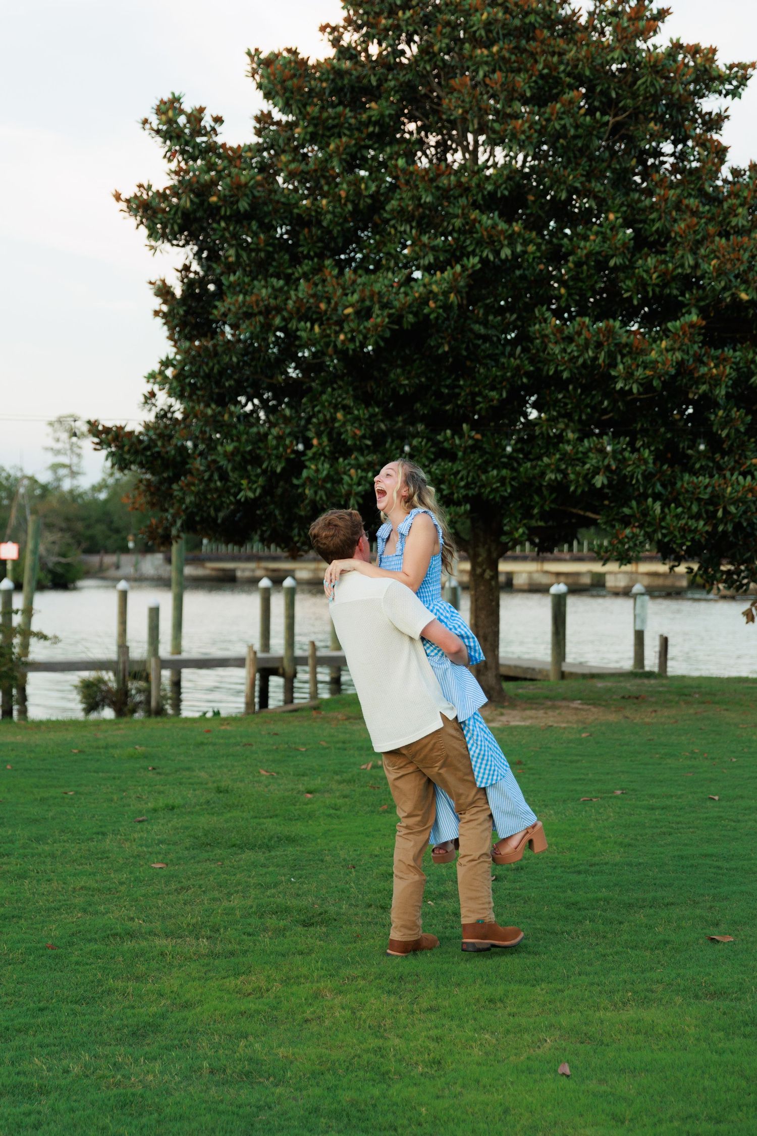 A man lifts a laughing woman in a blue dress on a green lawn near a waterfront with a large tree backdrop.