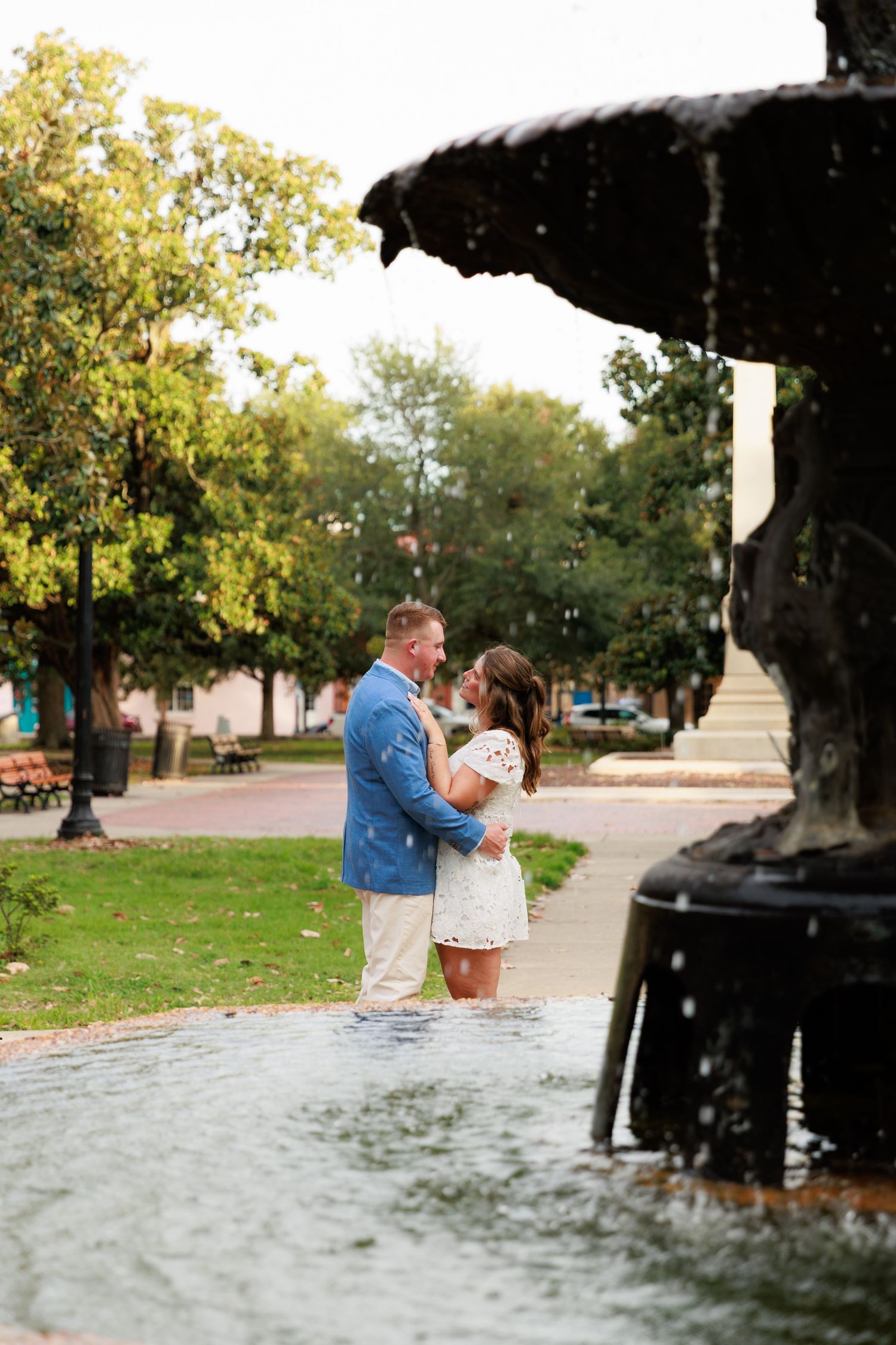 Couple embracing and gazing at each other beside a decorative fountain in a scenic outdoor park setting.