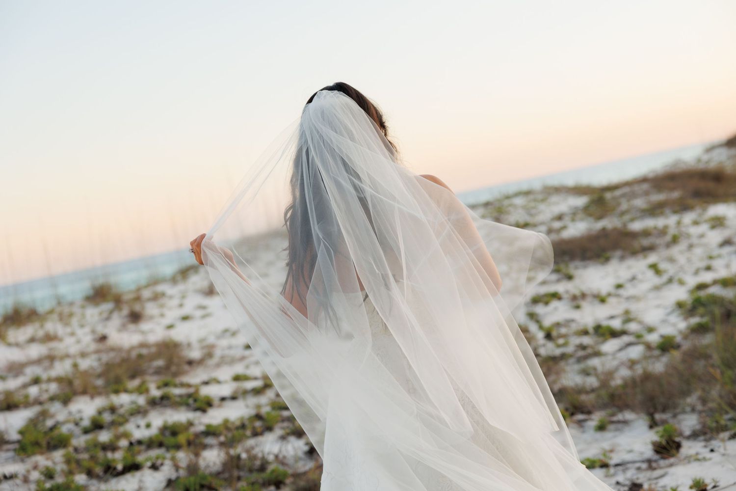 Bride in white wedding dress and veil standing on sandy beach at sunset, back facing camera.