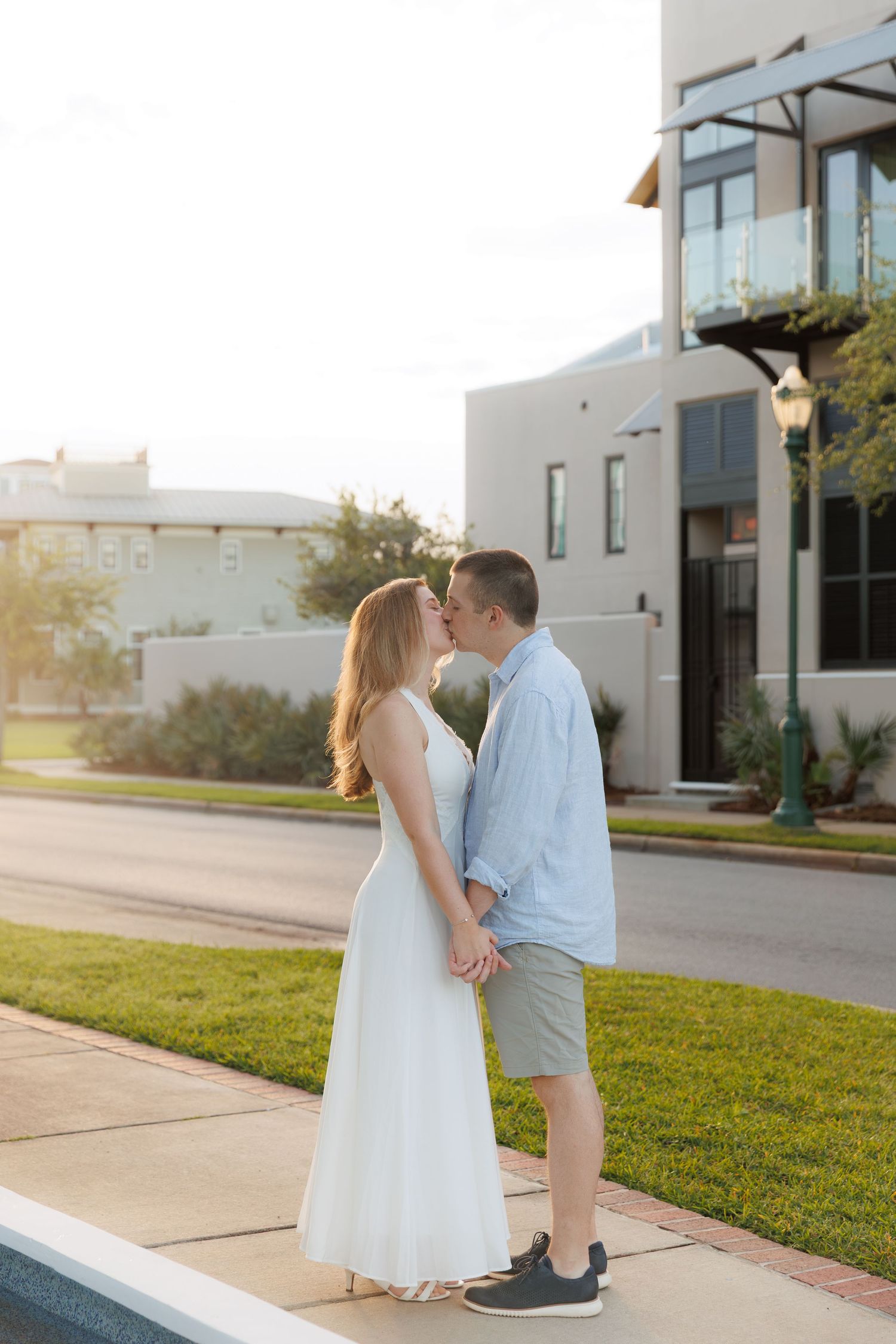Couple sharing a kiss while holding hands outdoors near a modern building at golden hour.