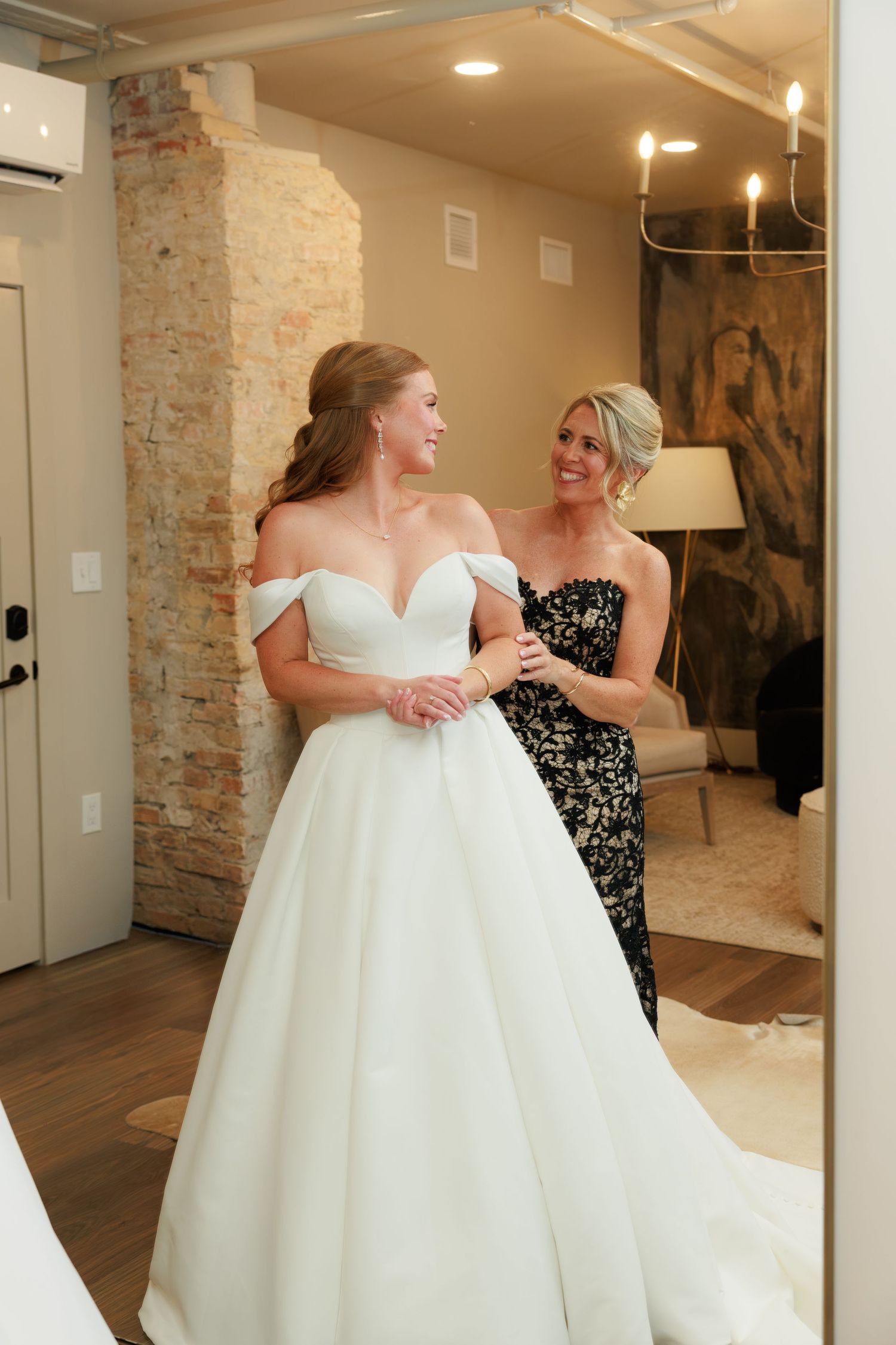 Bride in off-shoulder white ball gown with woman in black floral dress in elegant bridal boutique.