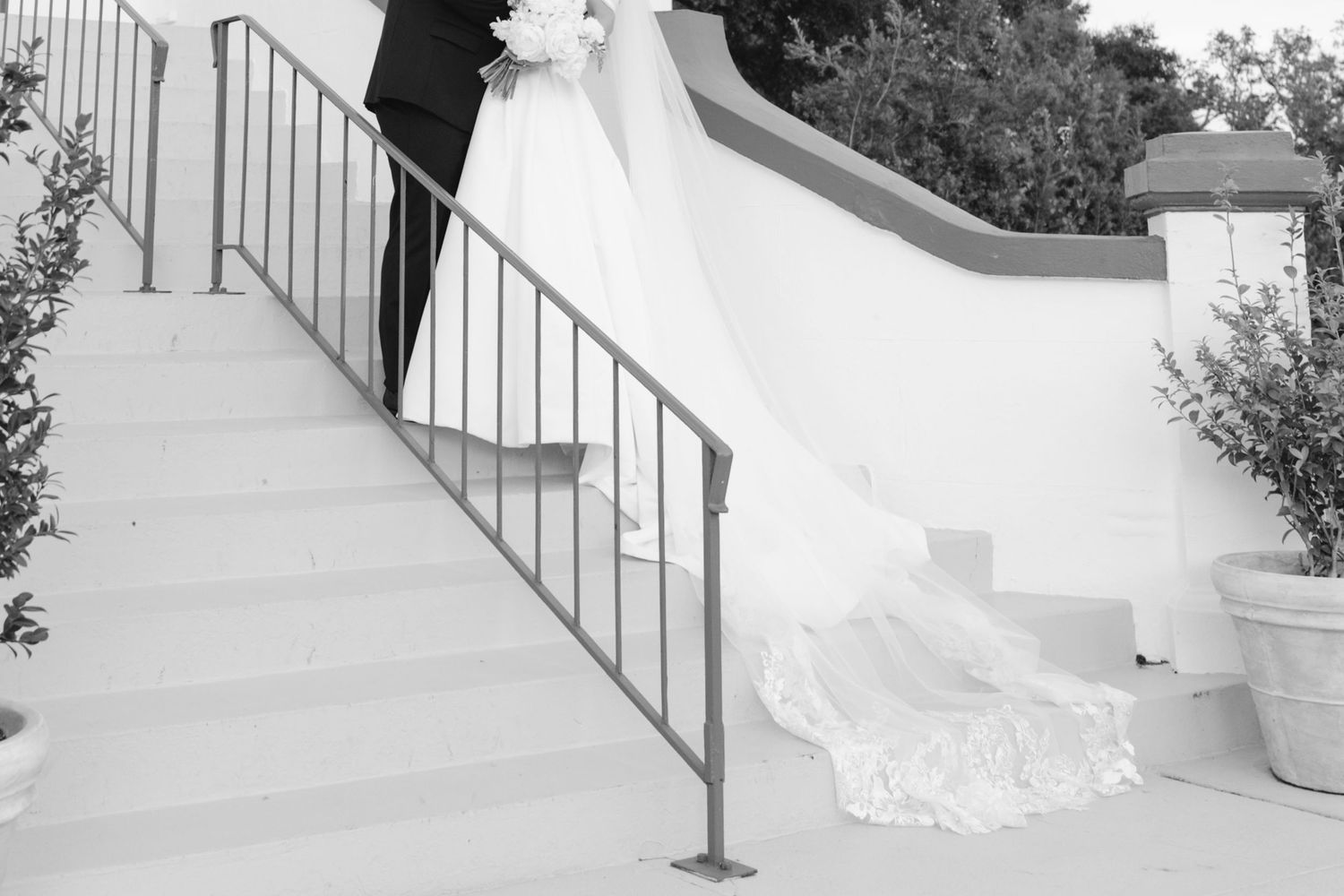 Black and white photo of a bride and groom walking up outdoor stairs, with wedding dress train flowing elegantly behind.