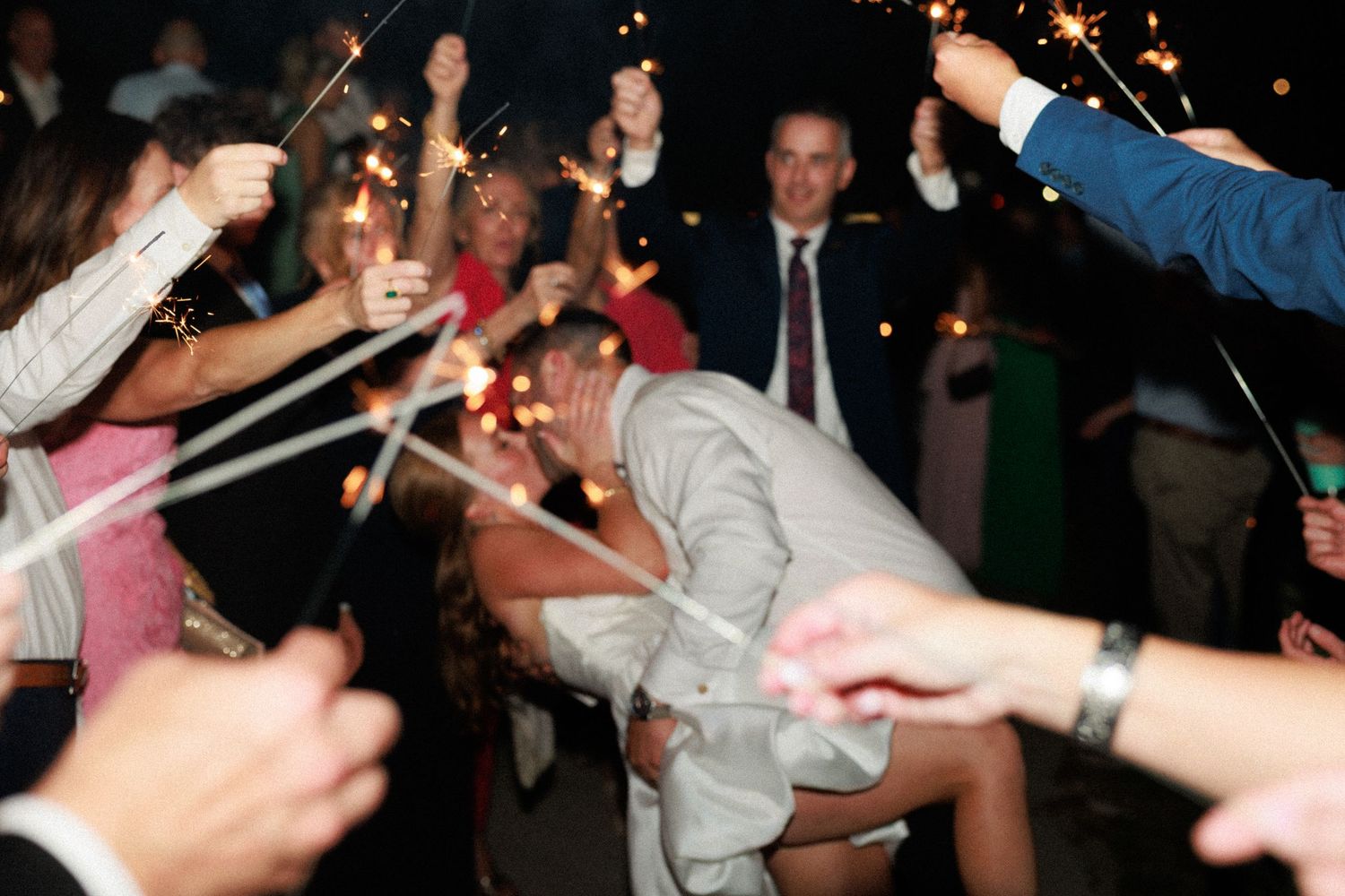 Wedding guests holding sparklers celebrate as couple shares a romantic kiss during festive sparkler send-off at night.