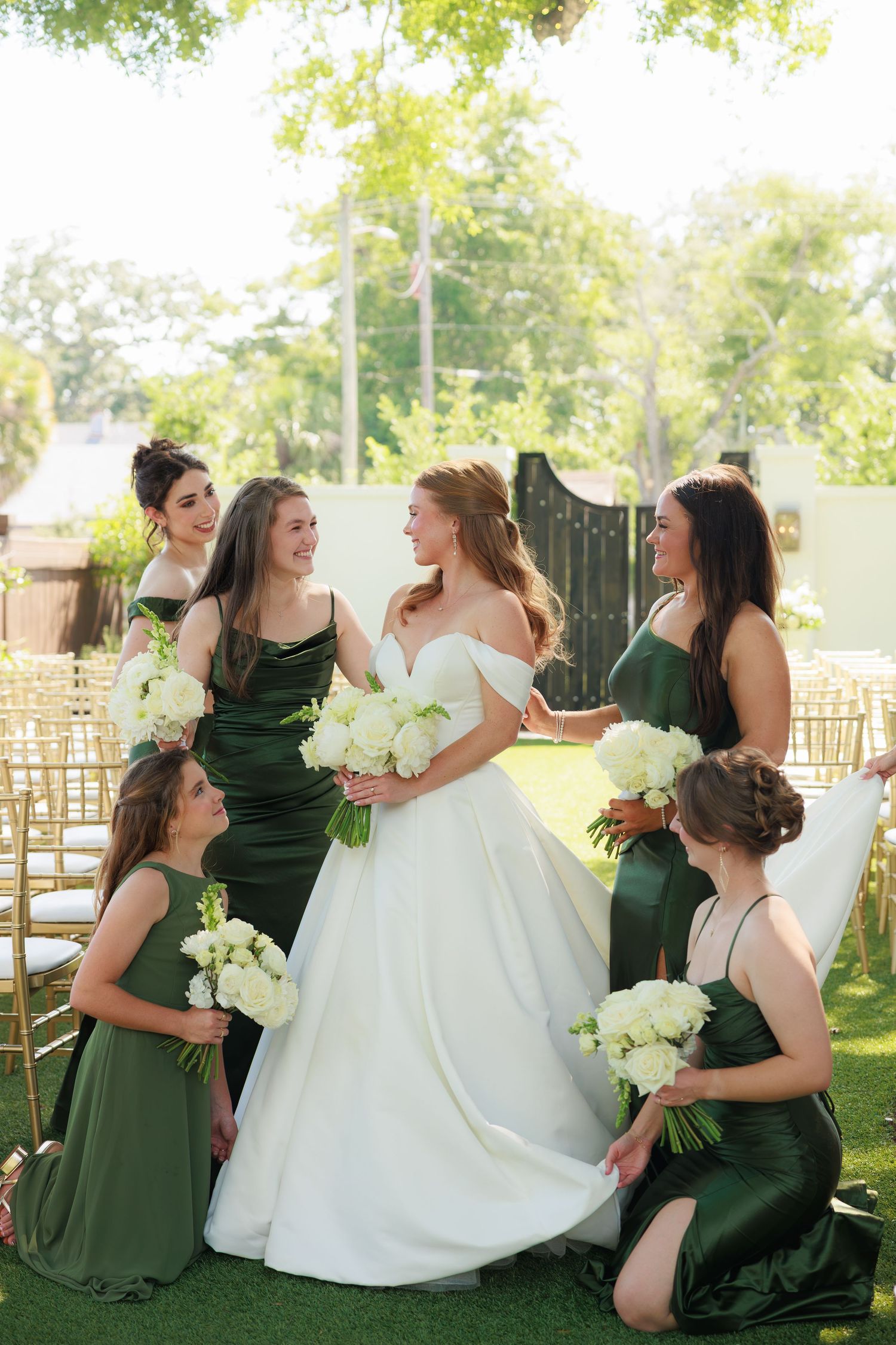 Bride in white gown surrounded by bridesmaids in emerald green dresses holding white bouquets at outdoor wedding ceremony.