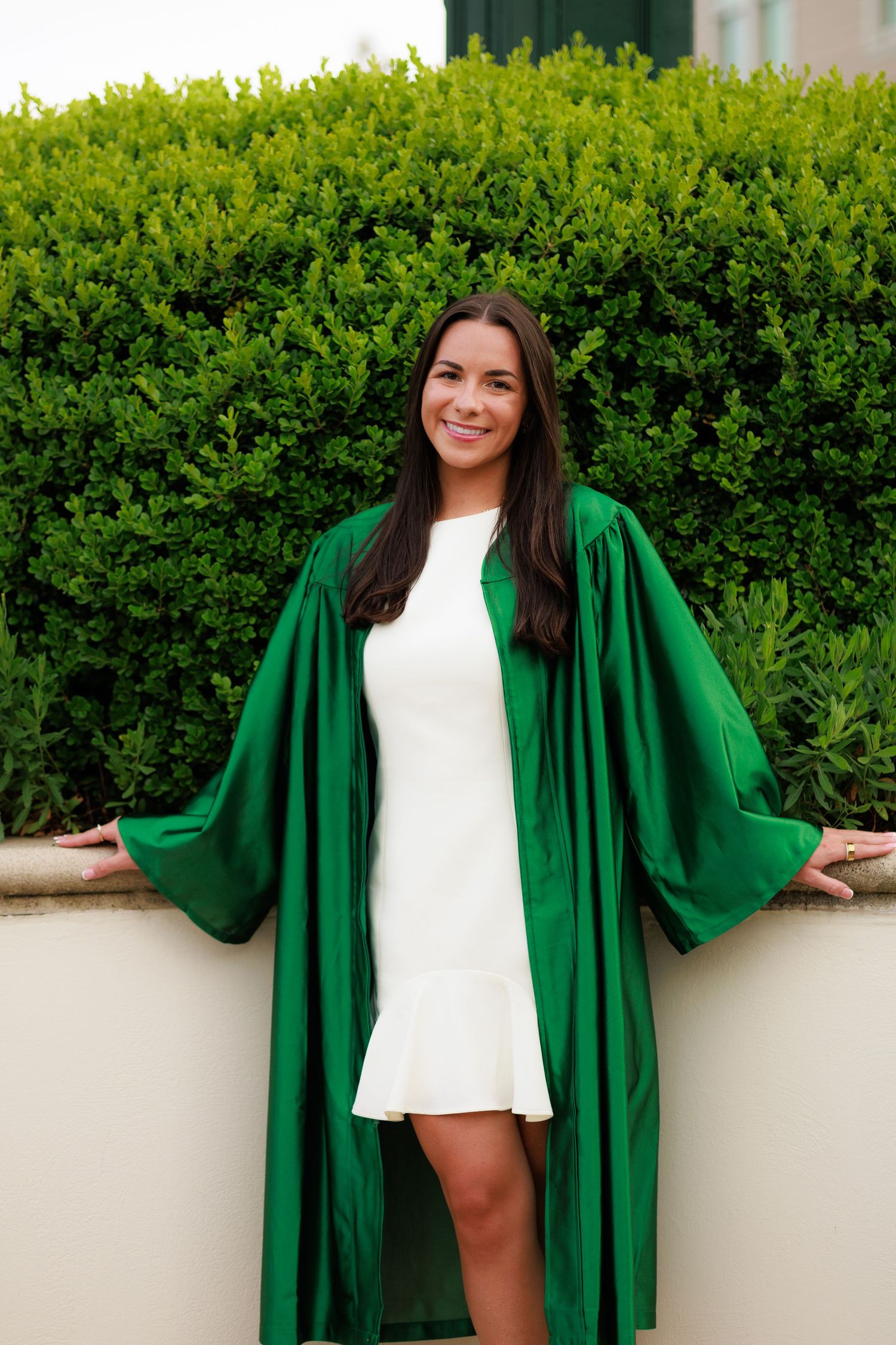Smiling young woman in green graduation gown and white dress posing in front of lush green hedges.