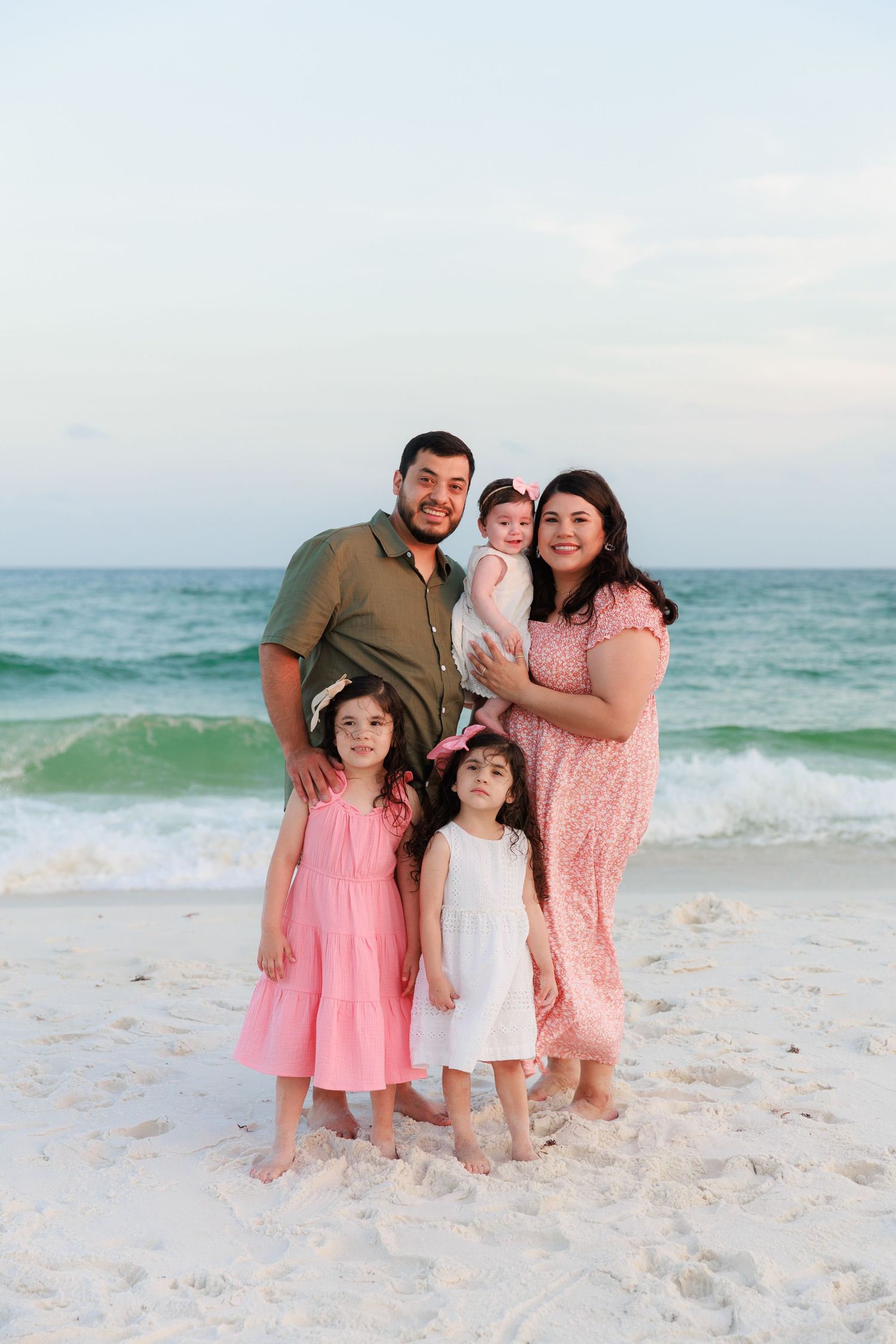 Family of five posing on a white sand beach with turquoise ocean waves, two girls in pink dresses and baby held by parents.