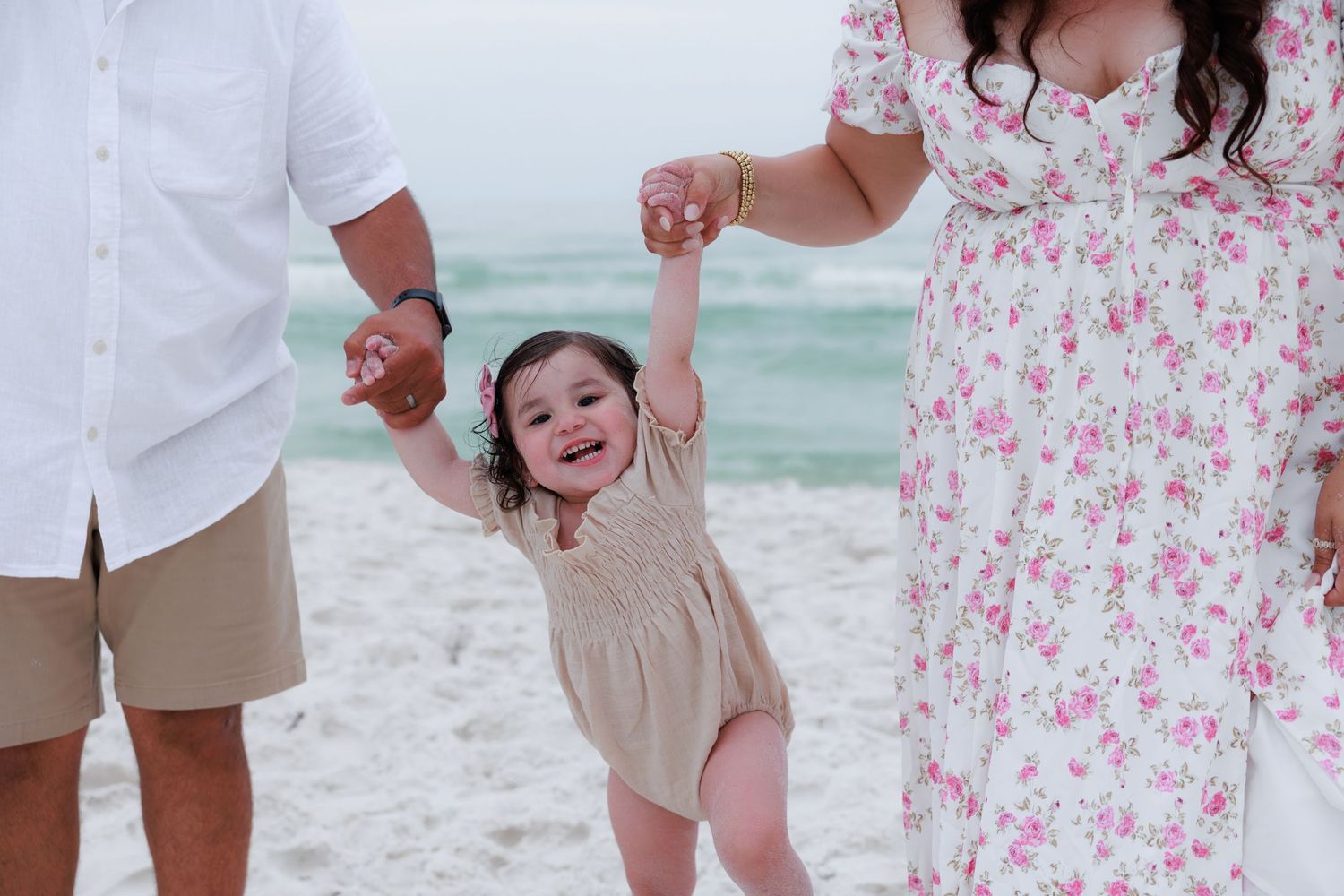 A joyful toddler holds parents hands on a sunny beach, wearing a beige romper, with turquoise waves in the background.