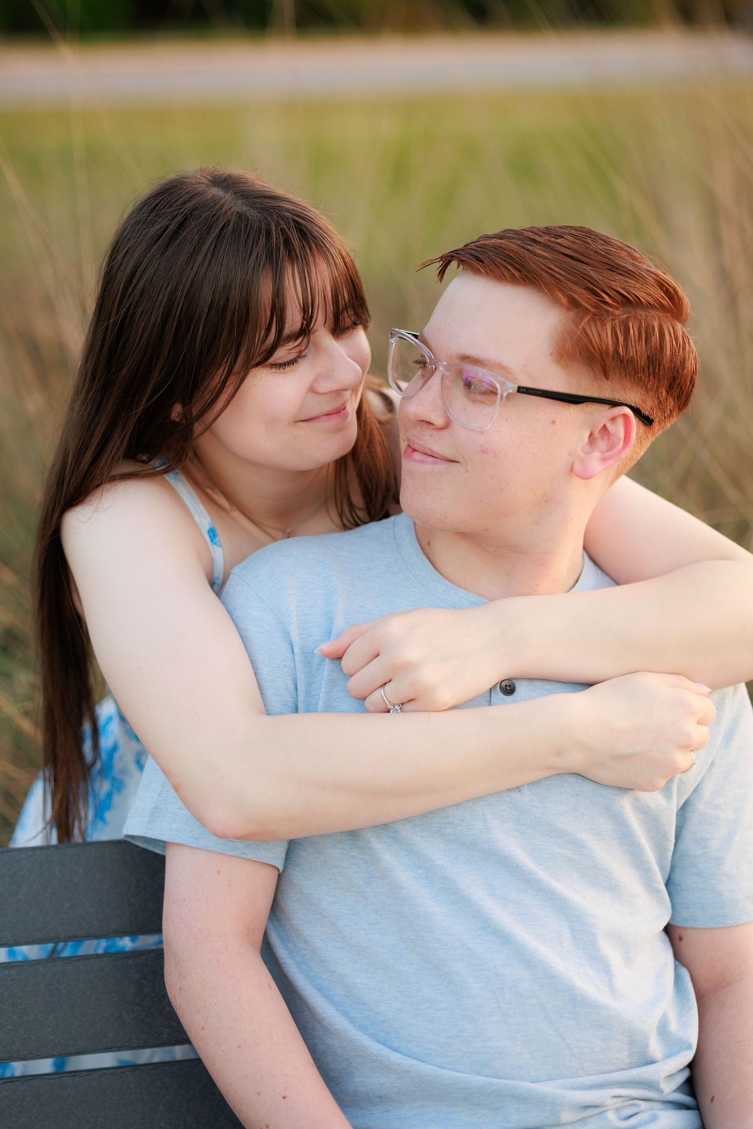 A young couple sitting on a bench outdoors, woman with dark hair embracing a red-haired man wearing glasses.