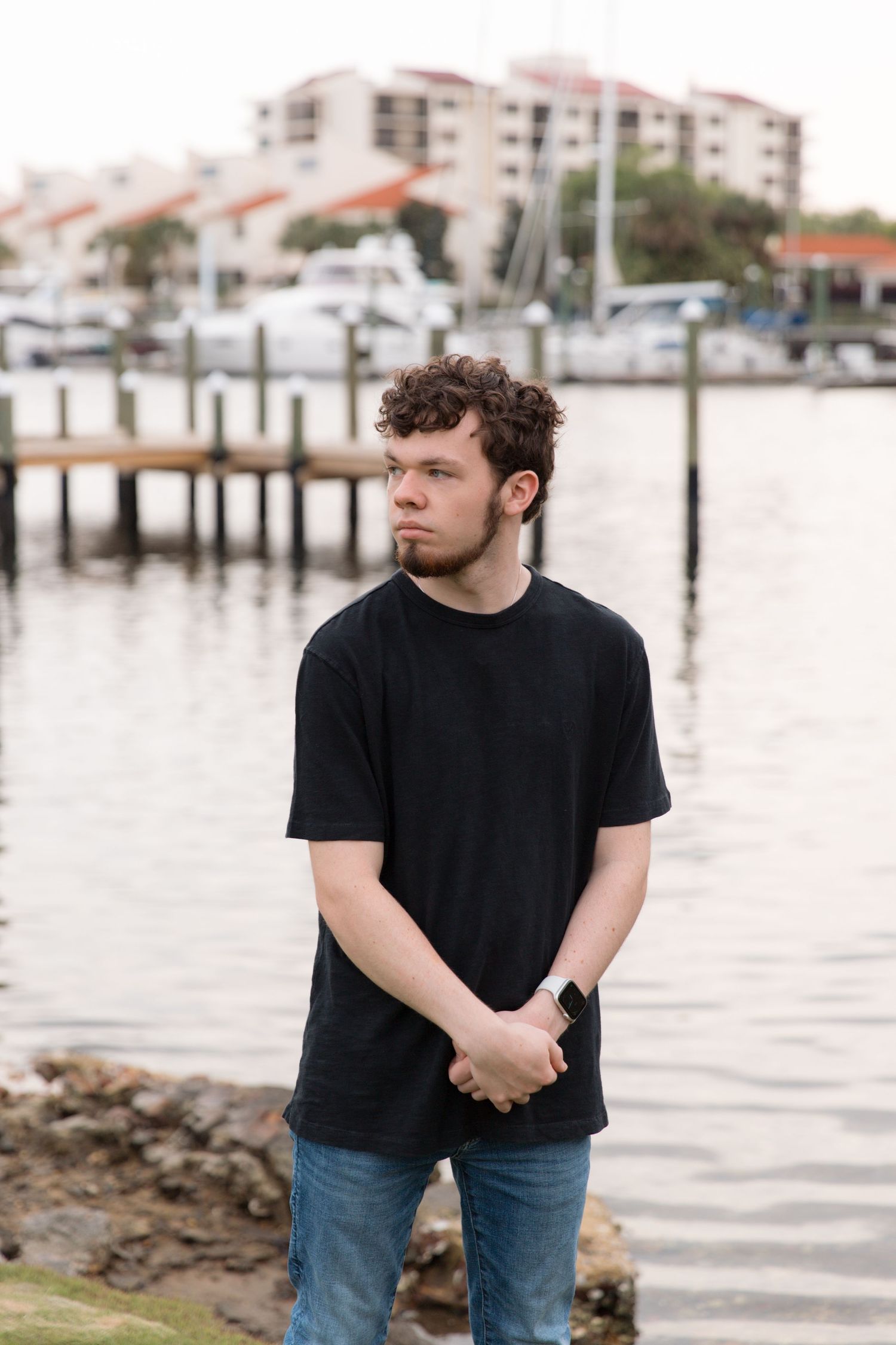 Standing by marina dock wearing black t-shirt with boats and buildings in background.