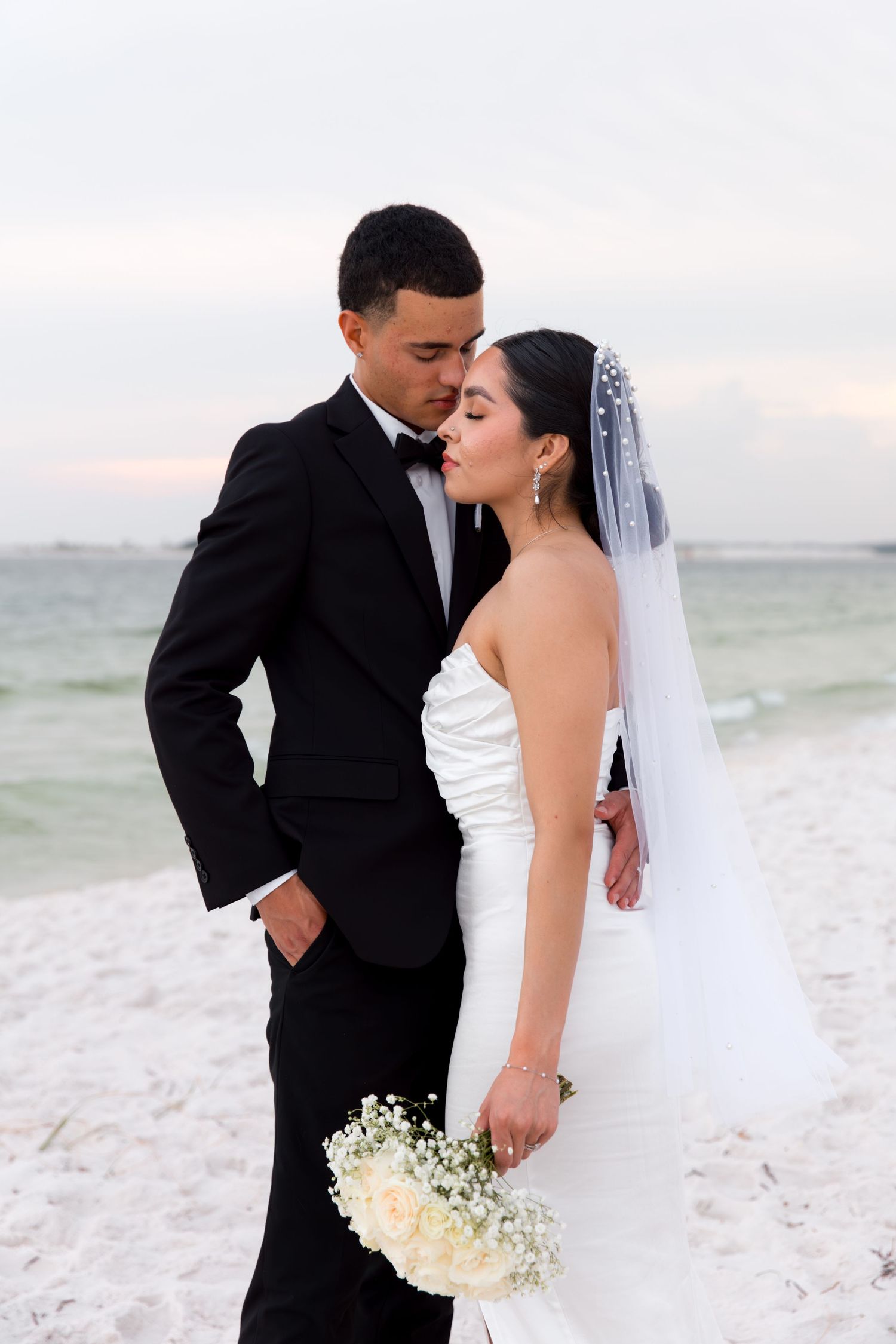 Elegant wedding couple poses on white sand beach at dusk.