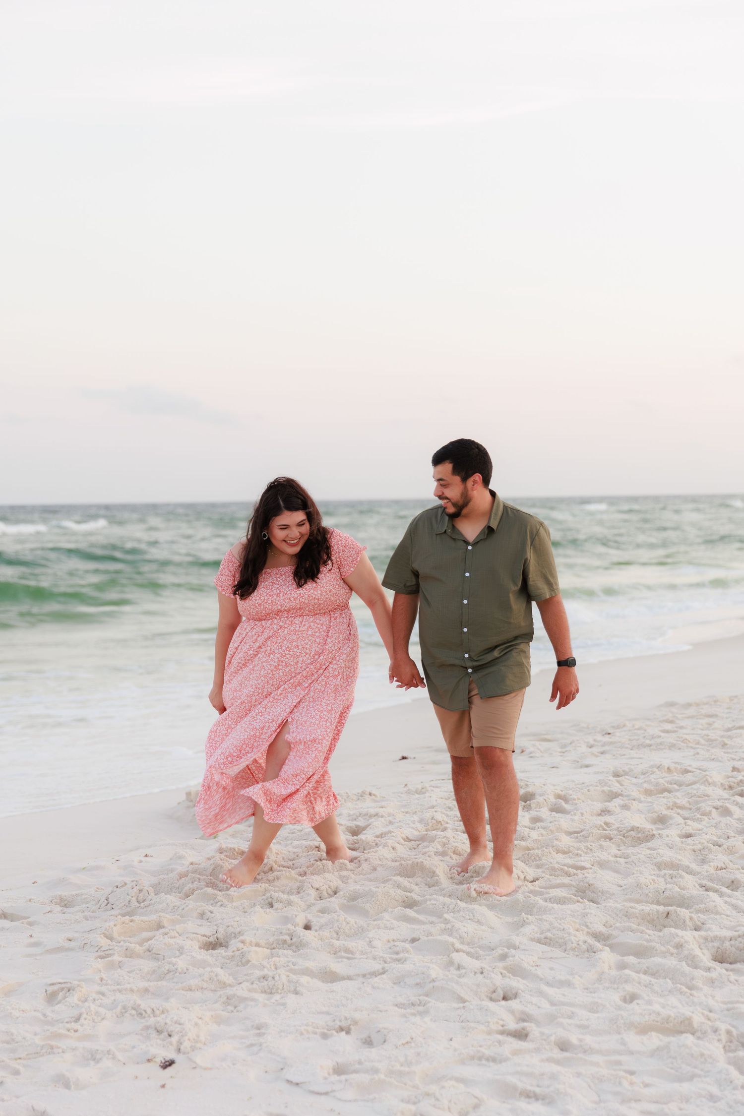 Couple holding hands walking on white sand beach at sunset, woman in pink floral dress, man in green shirt.