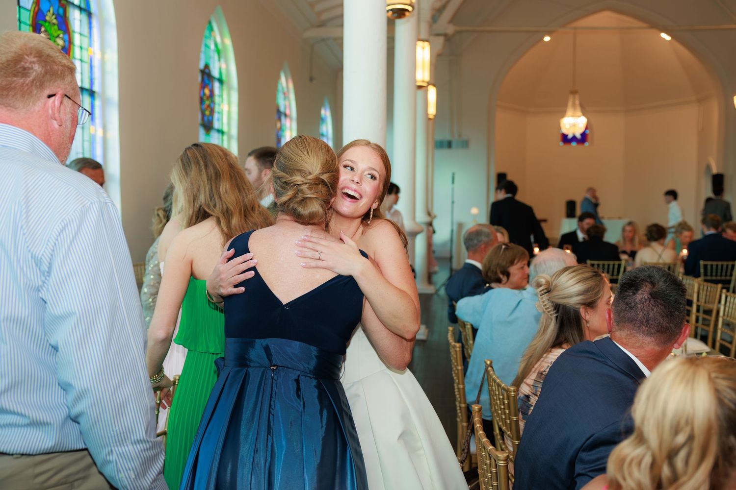 Bride hugging a guest in a navy dress at a wedding reception inside a beautiful chapel.