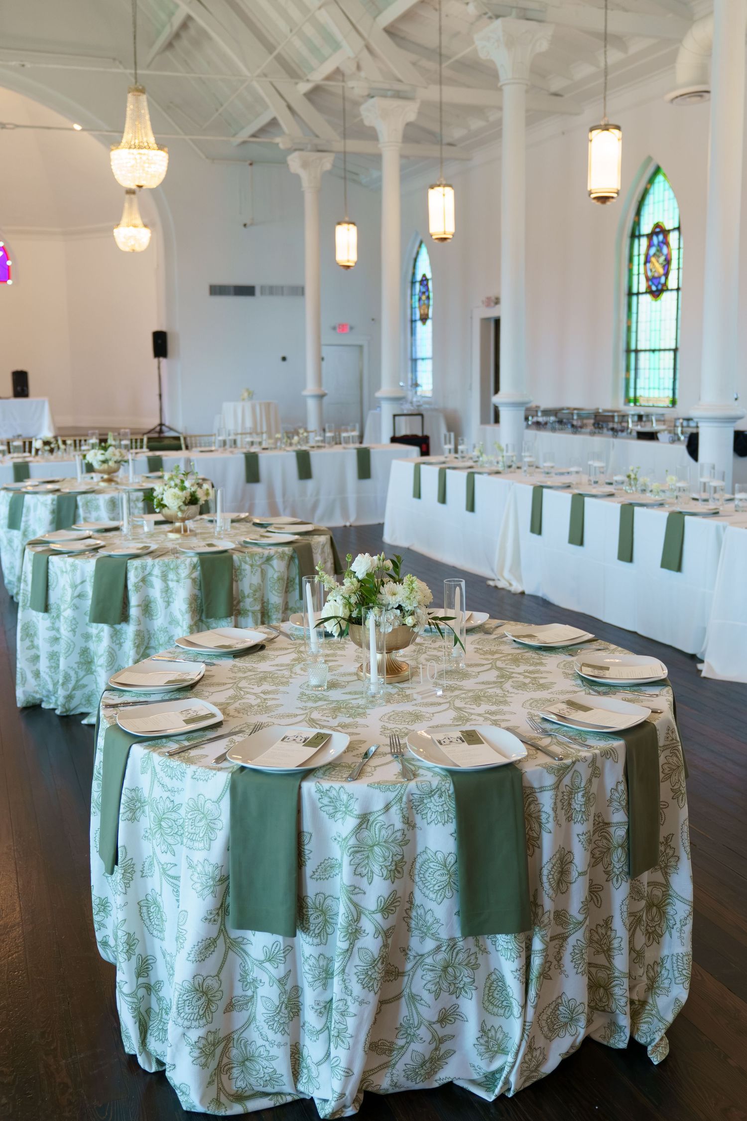 Elegant wedding reception hall with round tables featuring green and white floral linens, sage napkins, and stained glass windows.