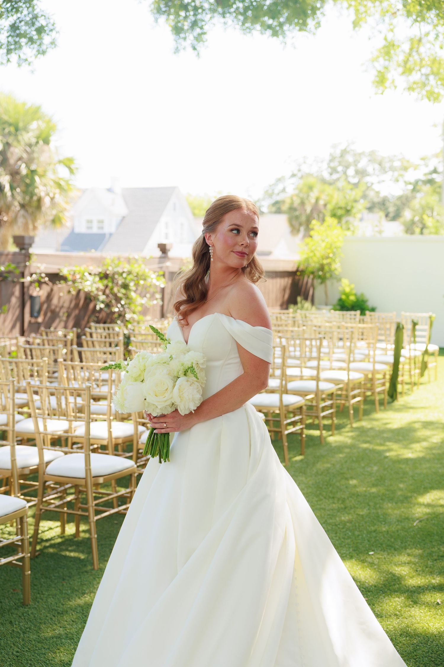 Bride in off-shoulder white ball gown holds white bouquet at outdoor wedding ceremony with gold chiavari chairs.