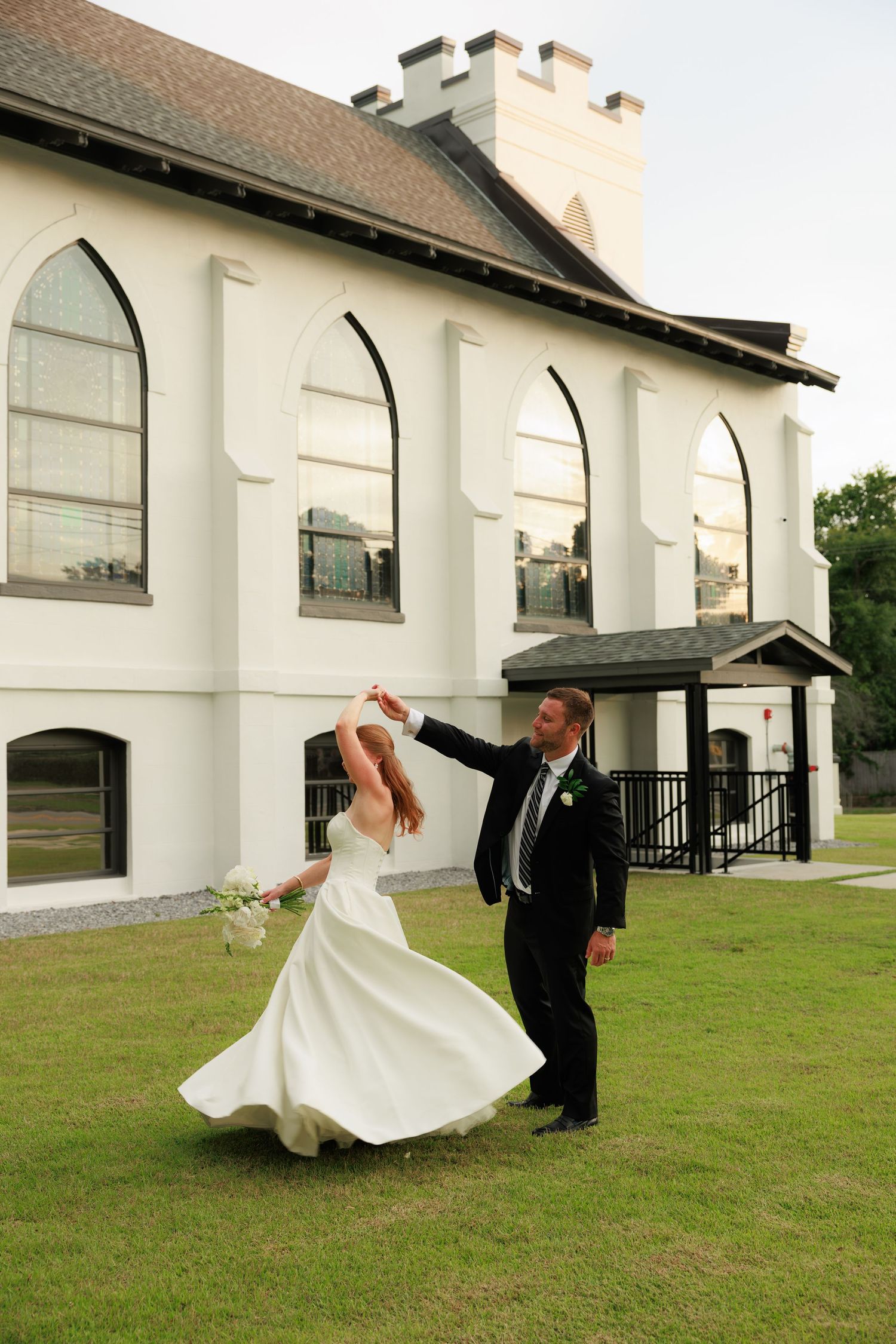 Groom twirls bride in flowing white gown on church lawn, her hair and dress spinning gracefully.