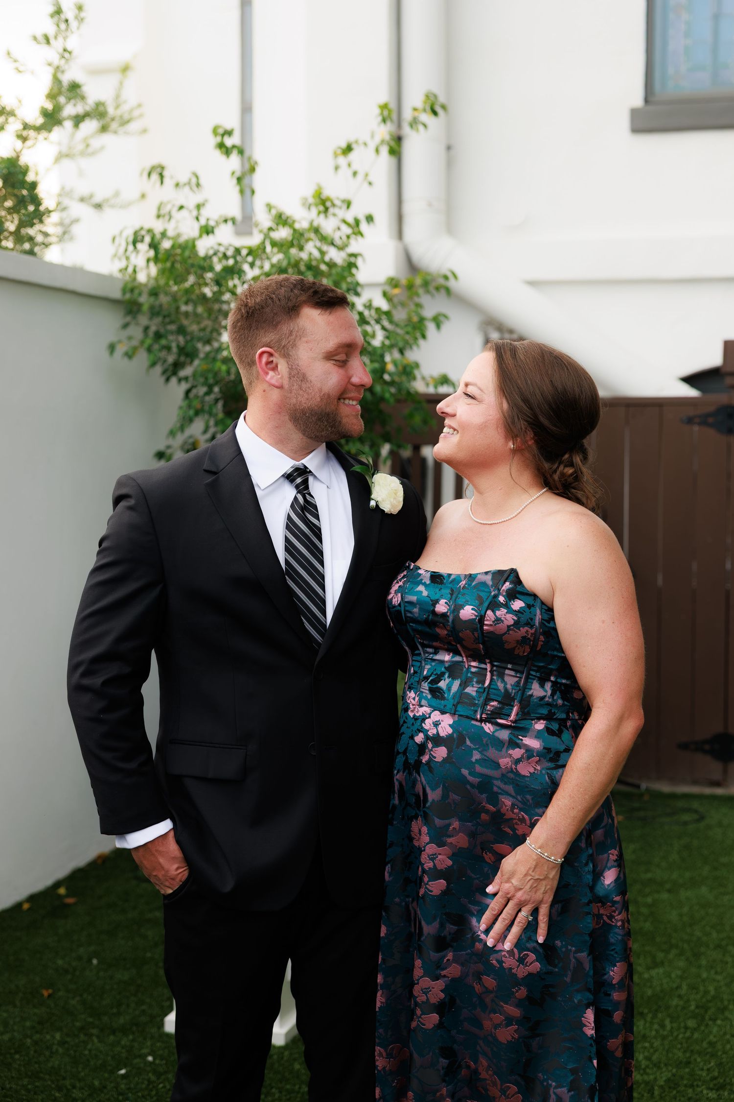 A man in a black suit and a woman in a floral teal dress smile at each other at an outdoor wedding event.