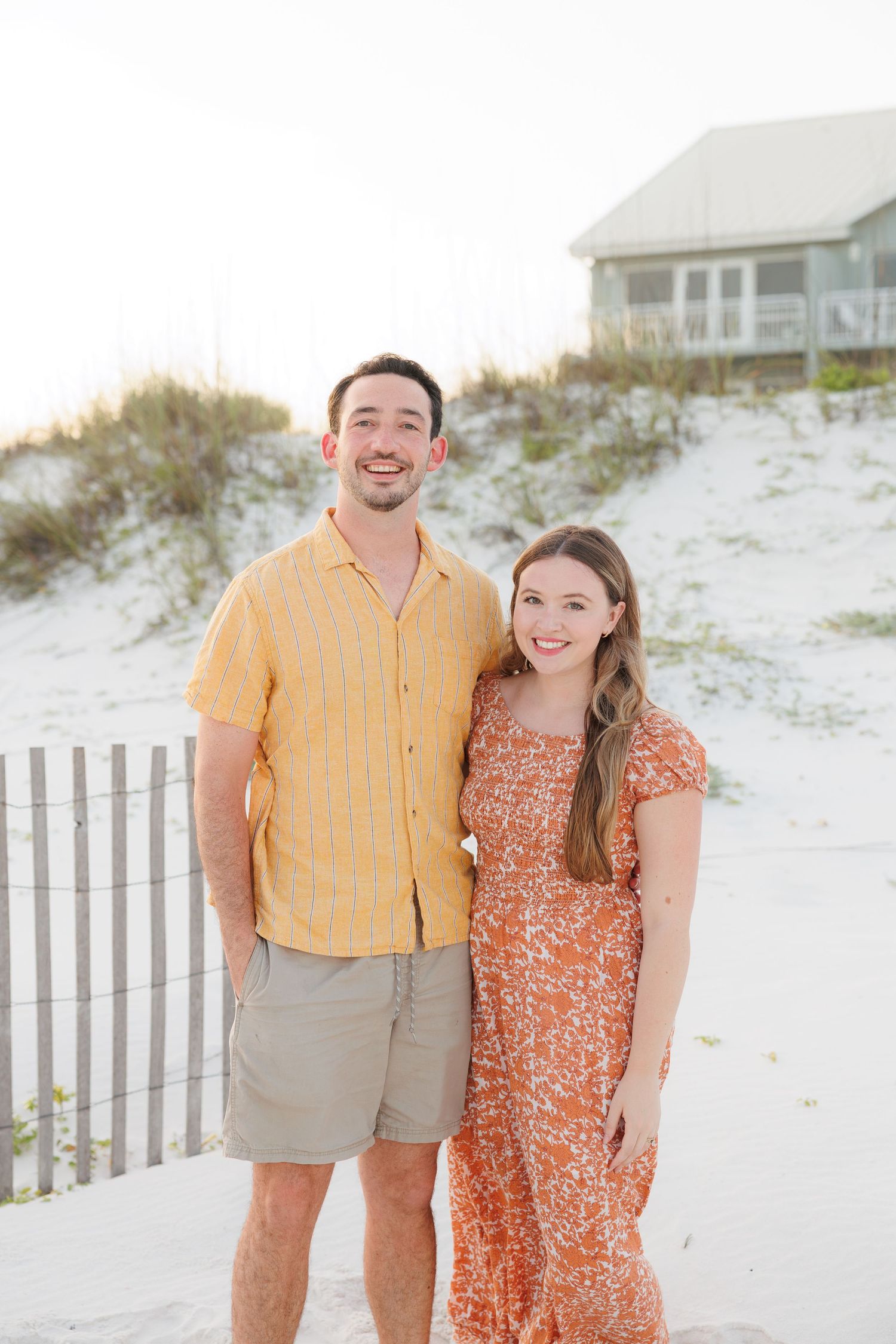 Smiling couple posing on a white sand beach, man in yellow shirt and woman in orange floral dress.