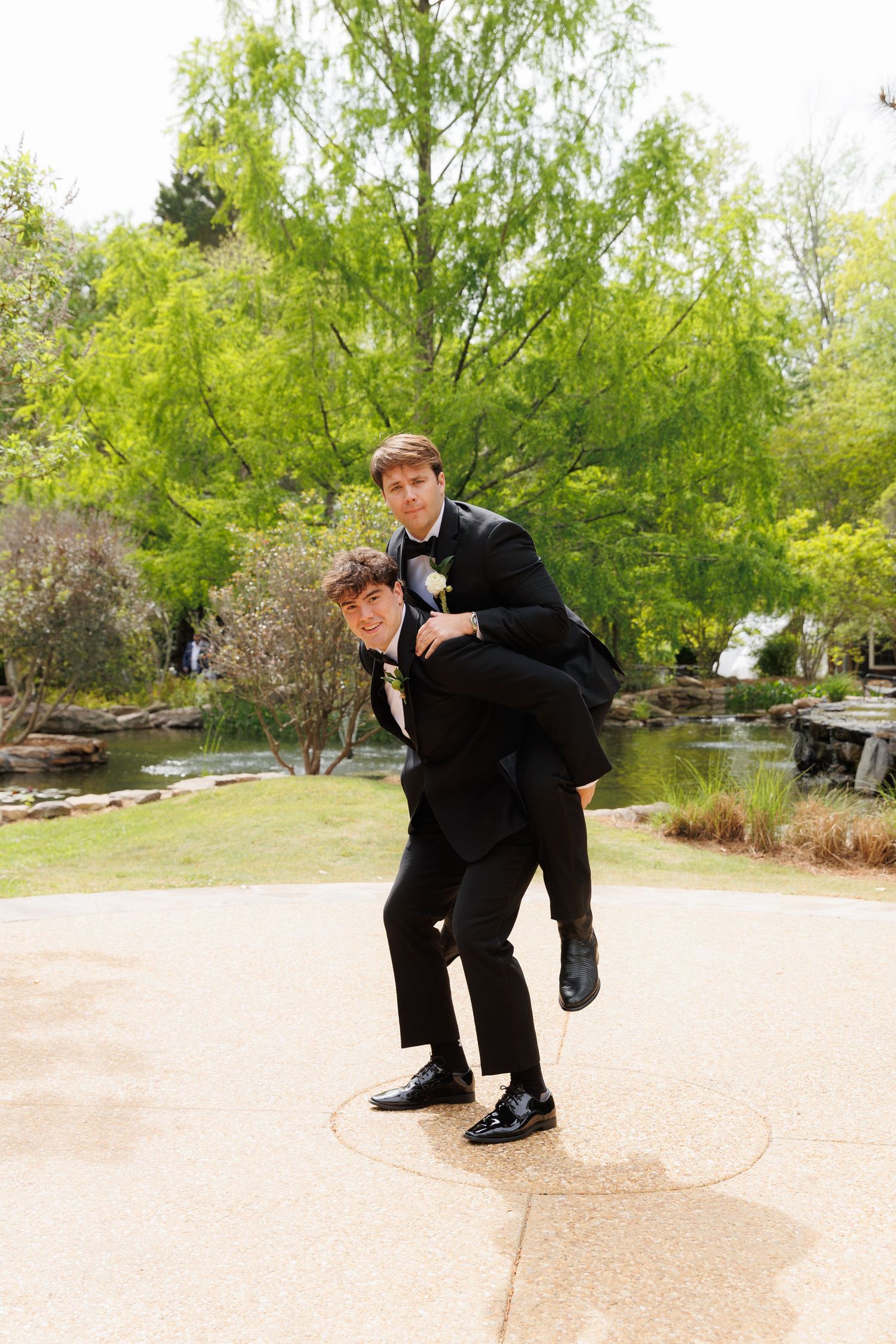 Two groomsmen in black tuxedos playfully posing with piggyback ride in an outdoor garden setting.