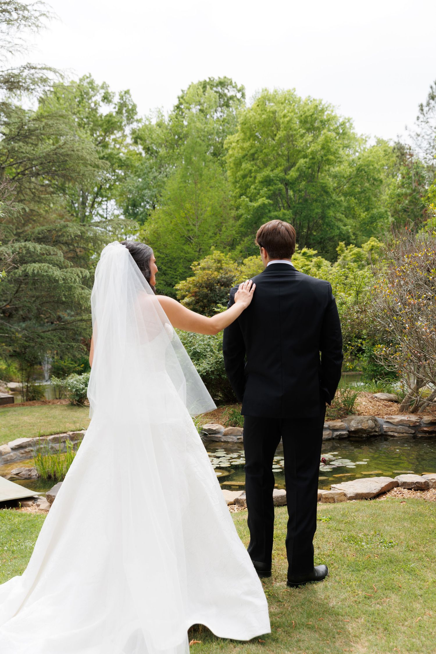 Bride in white gown with veil touches groom's back as couple stands before garden pond.