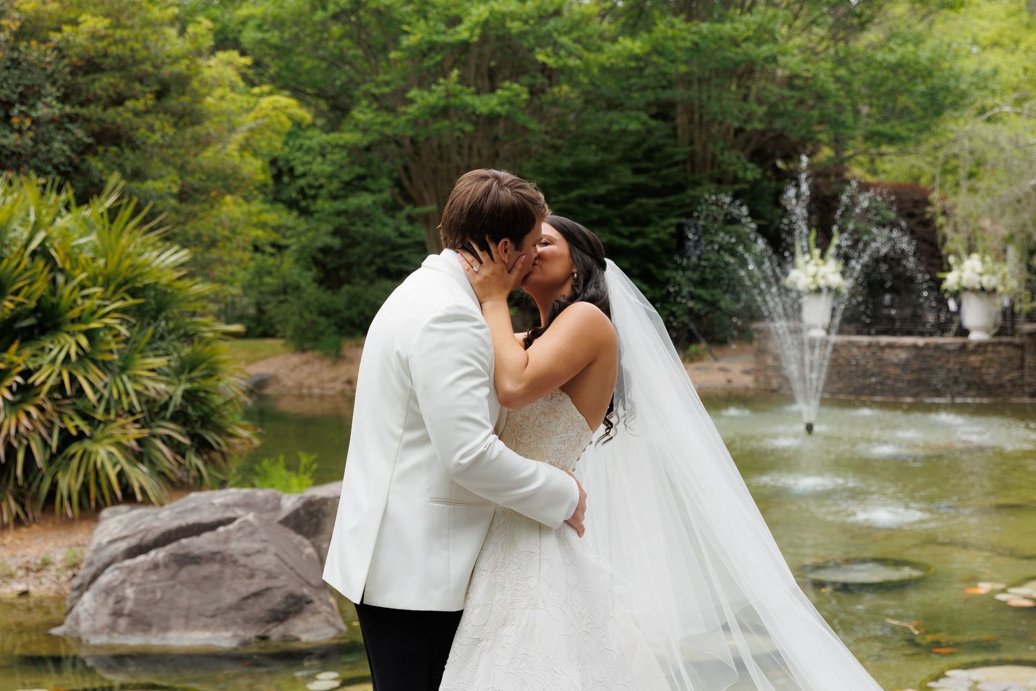 Bride and groom share a kiss near a garden fountain surrounded by lush greenery.