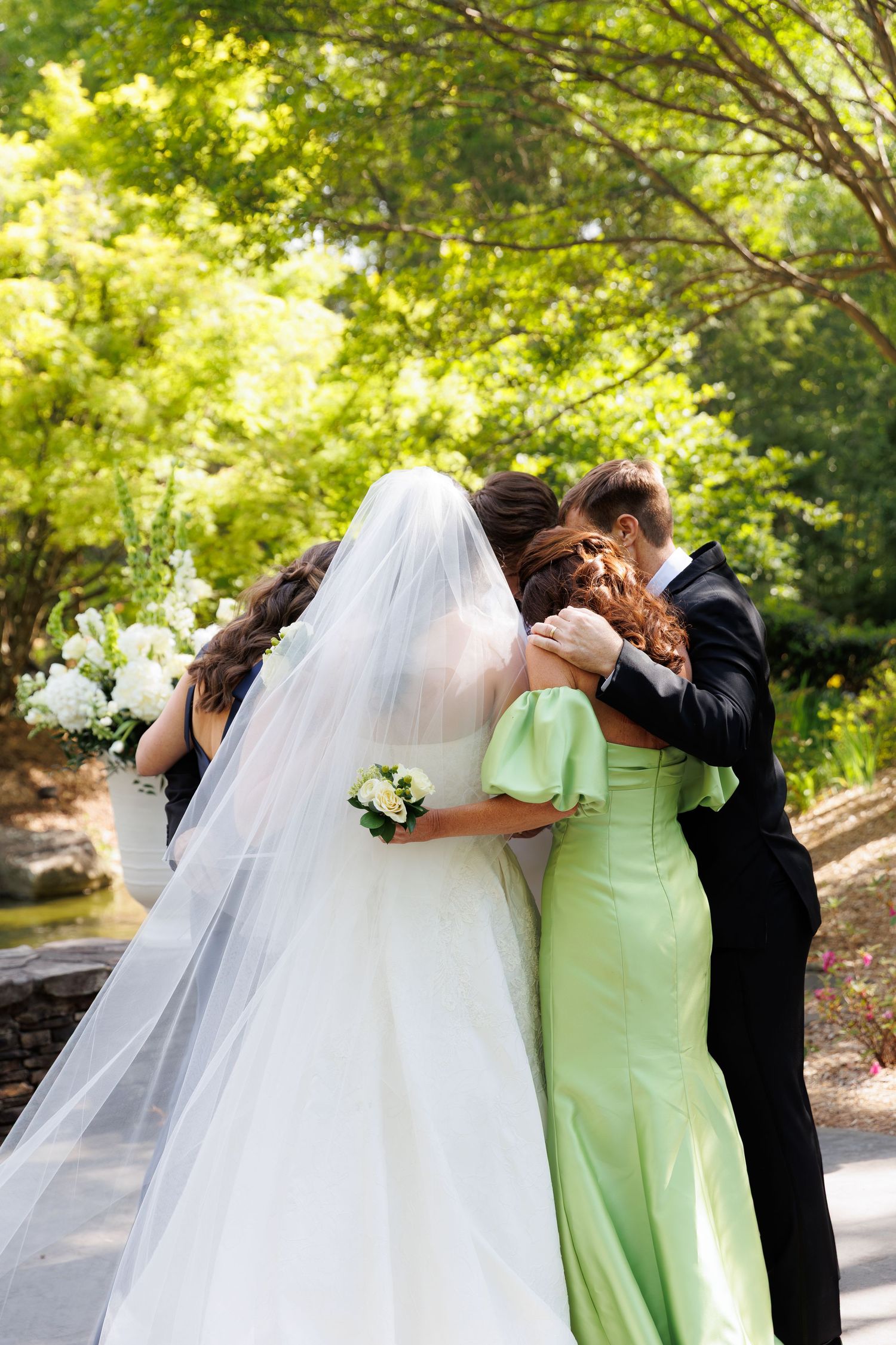 Bride in white gown and veil shares emotional hug with bridesmaid in green dress and groom outdoors.