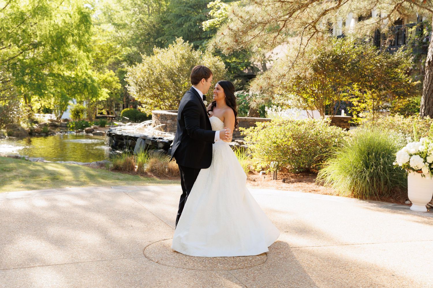 Bride and groom share a romantic moment in a lush garden with a pond, bride in a white ballgown.