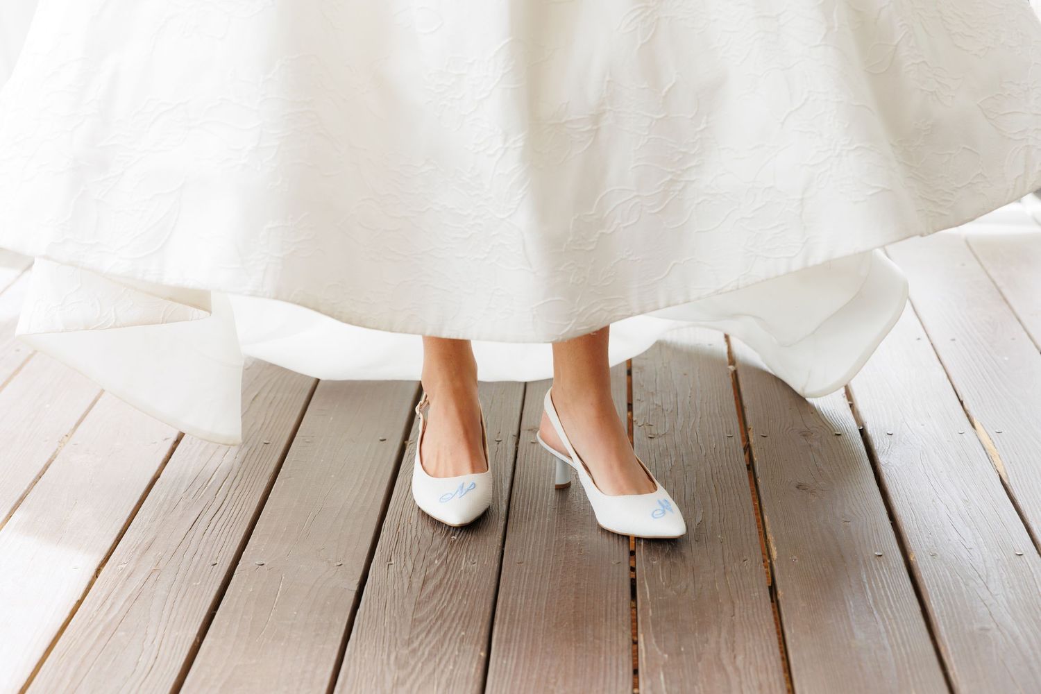 Close-up of a bride wearing white heeled wedding shoes and a flowing white bridal gown on a wooden floor.