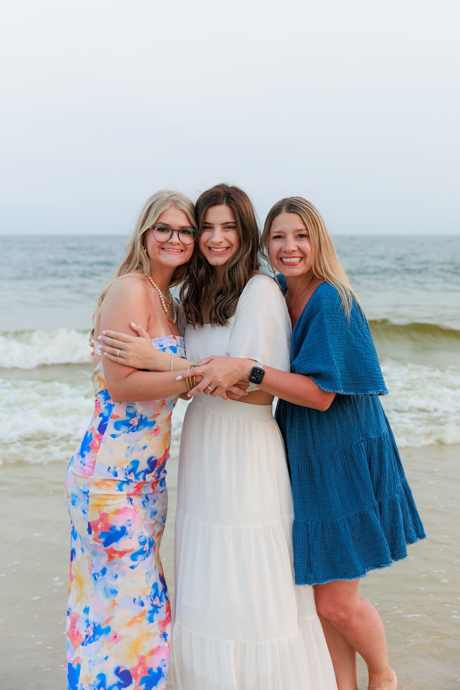 Three women smiling and hugging on a beach, wearing colorful, white, and blue dresses with ocean waves behind them.