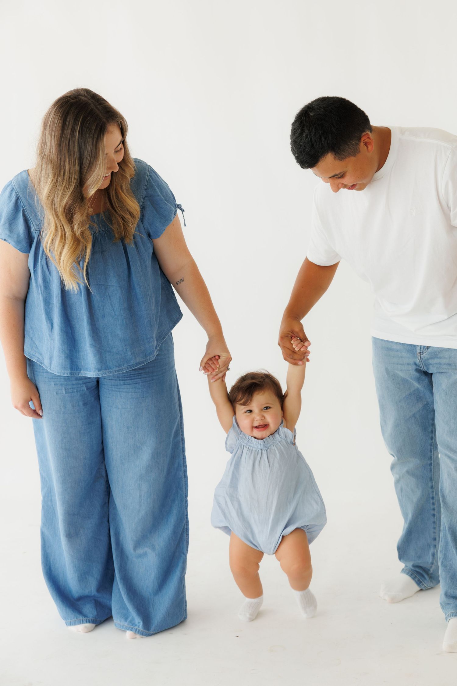 Happy parents help their baby girl take her first steps in a bright white studio, all dressed in blue denim outfits.
