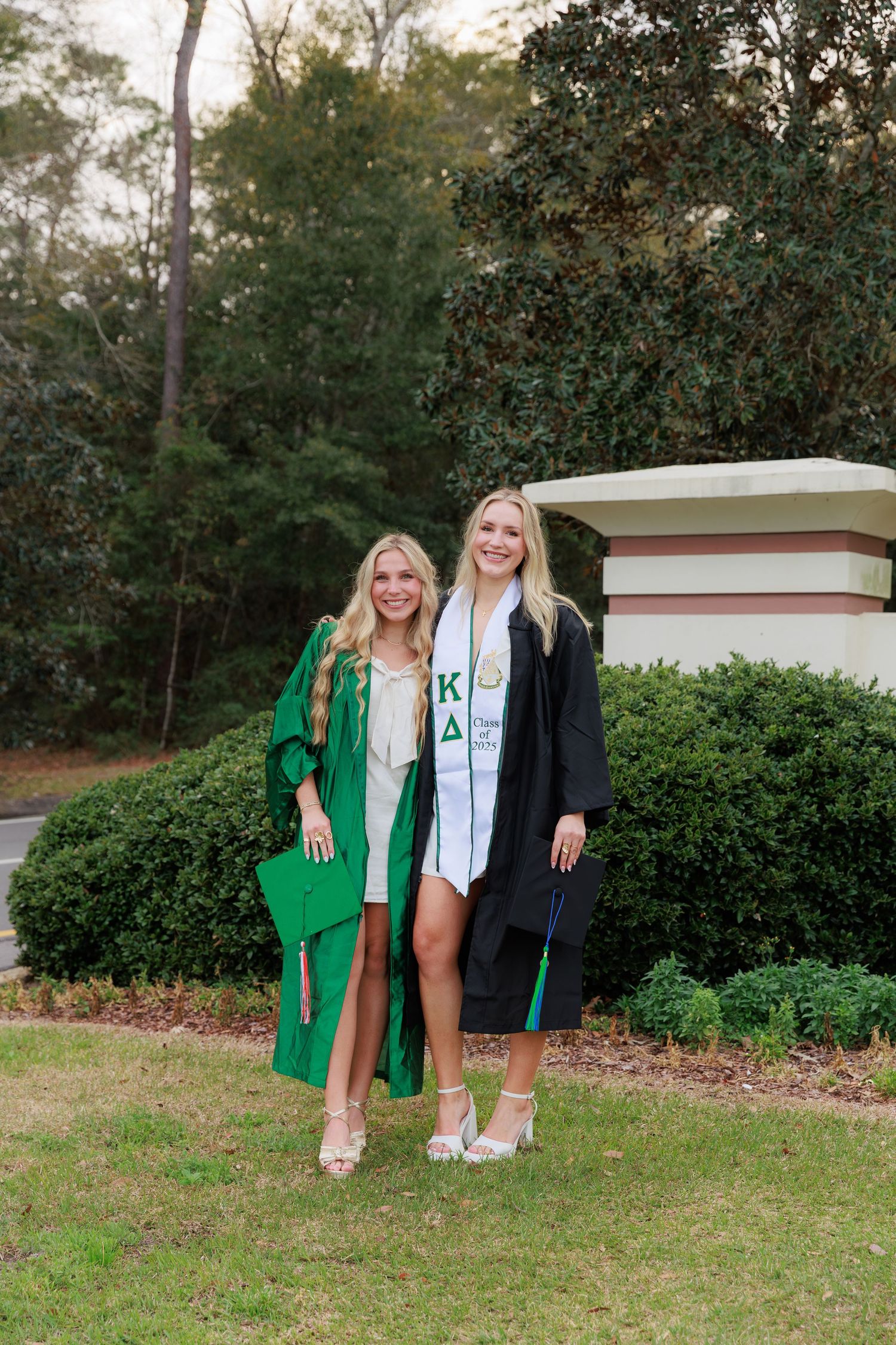 Two smiling blonde graduates in green and black caps and gowns posing outdoors on campus.