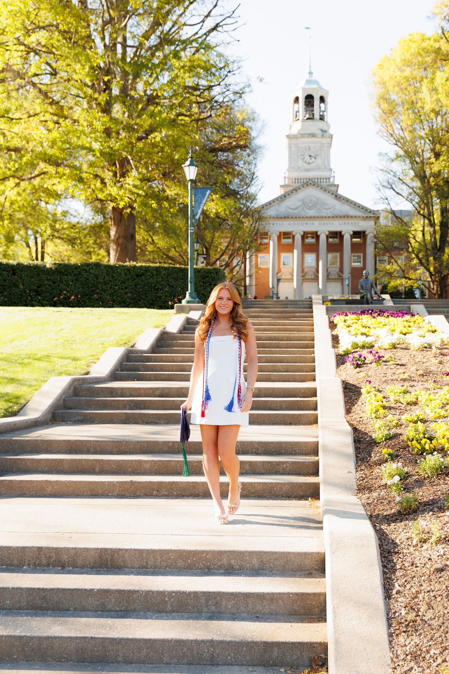 Young woman in white dress descending college campus steps with historic building and blooming flowers in background.