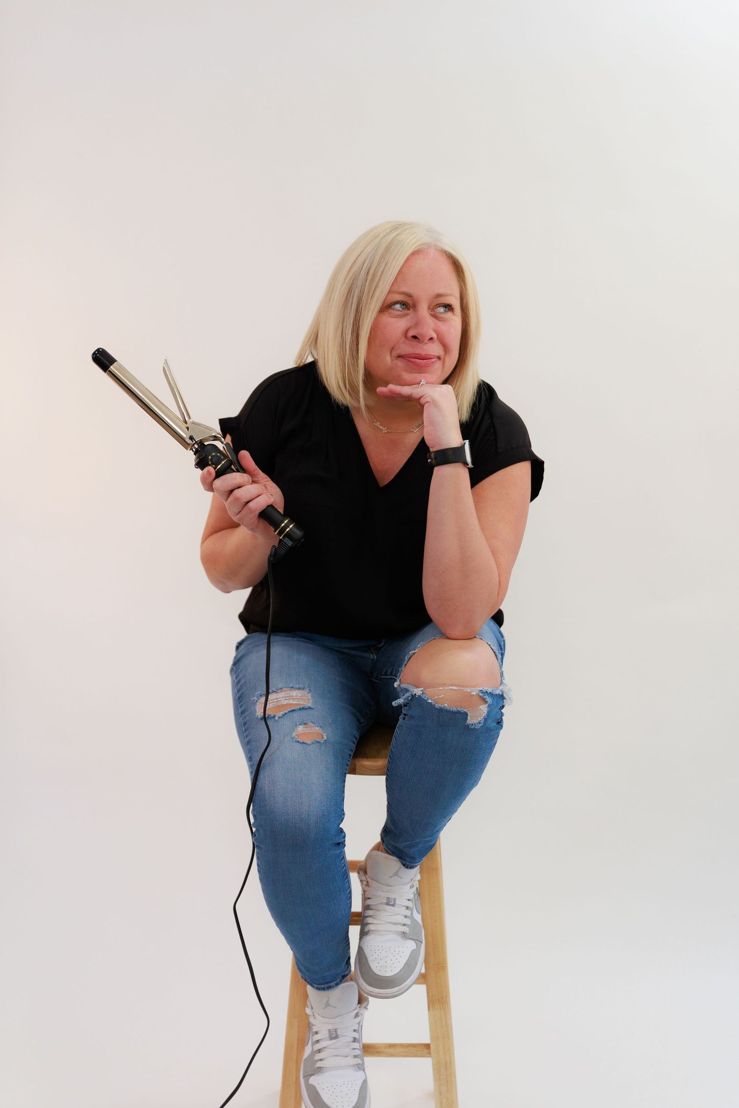 Casual portrait on wooden stool holding styling tools against white backdrop.
