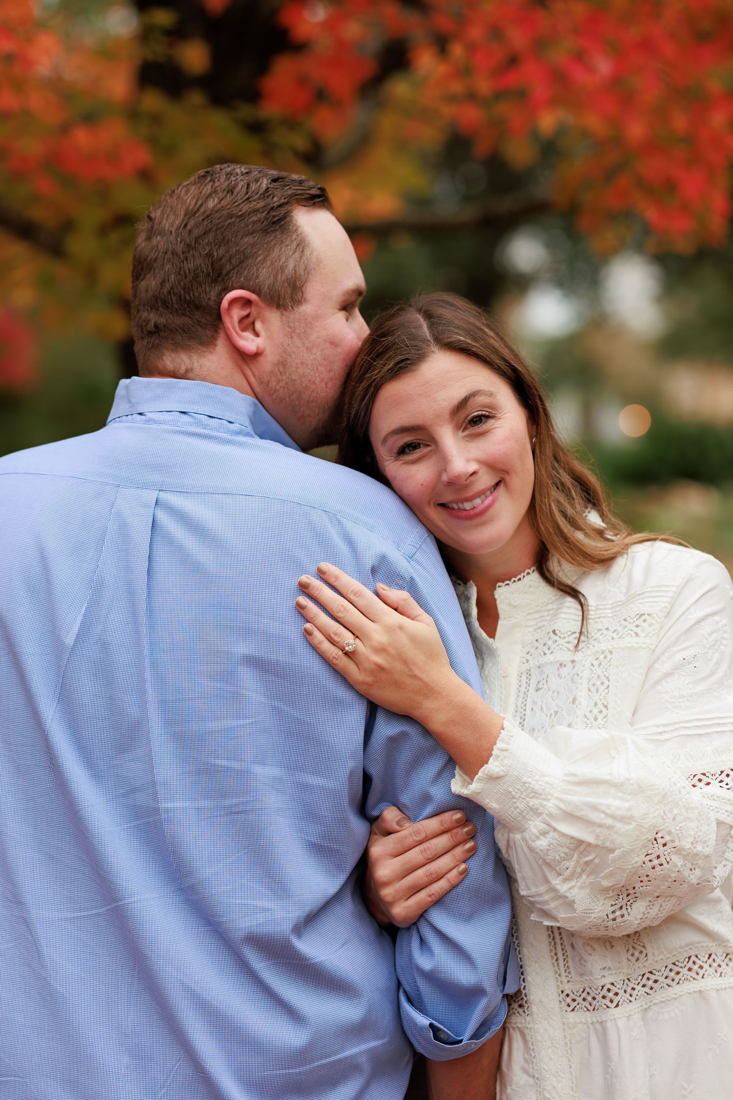 A couple embraces outdoors in autumn with vibrant red fall foliage in the background.