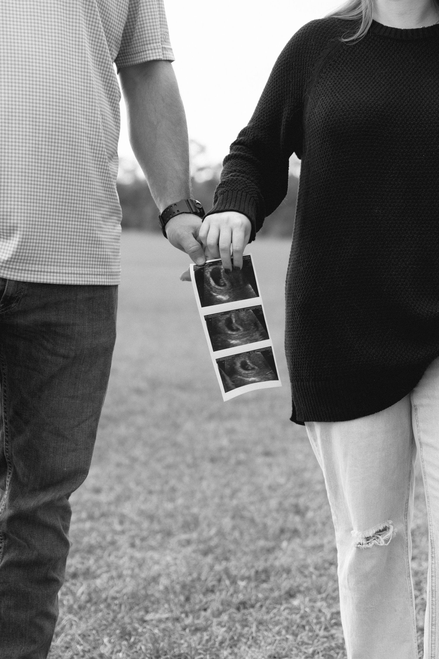 Black and white photo of couple holding ultrasound photos while standing together outdoors on grass.