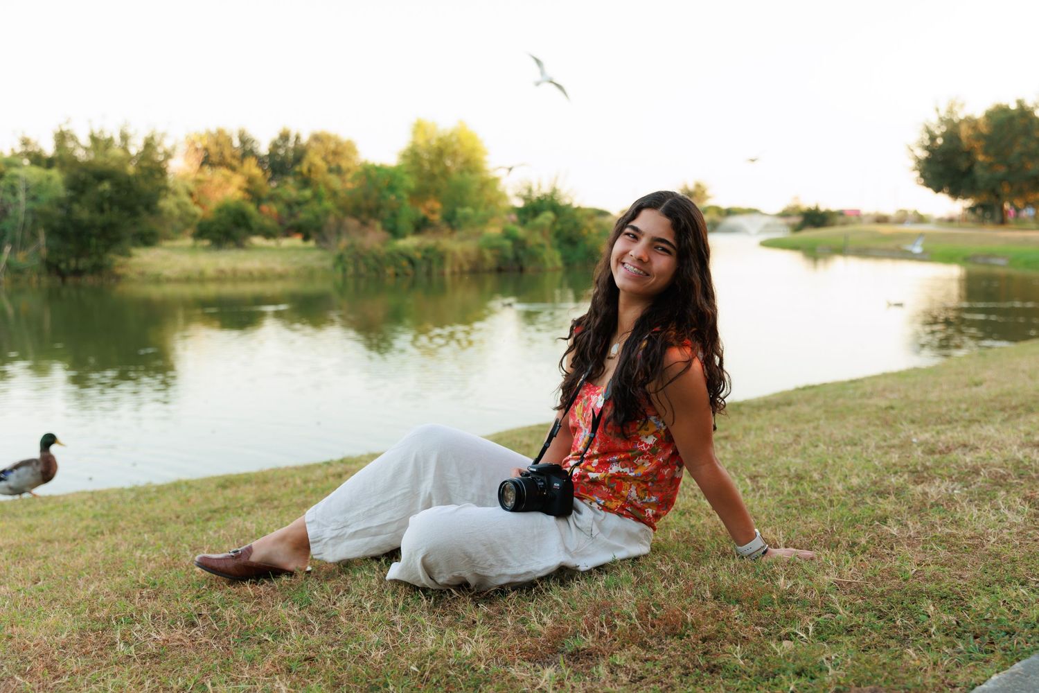 Woman in earth tones relaxing by tranquil lake on grassy shore during golden hour.